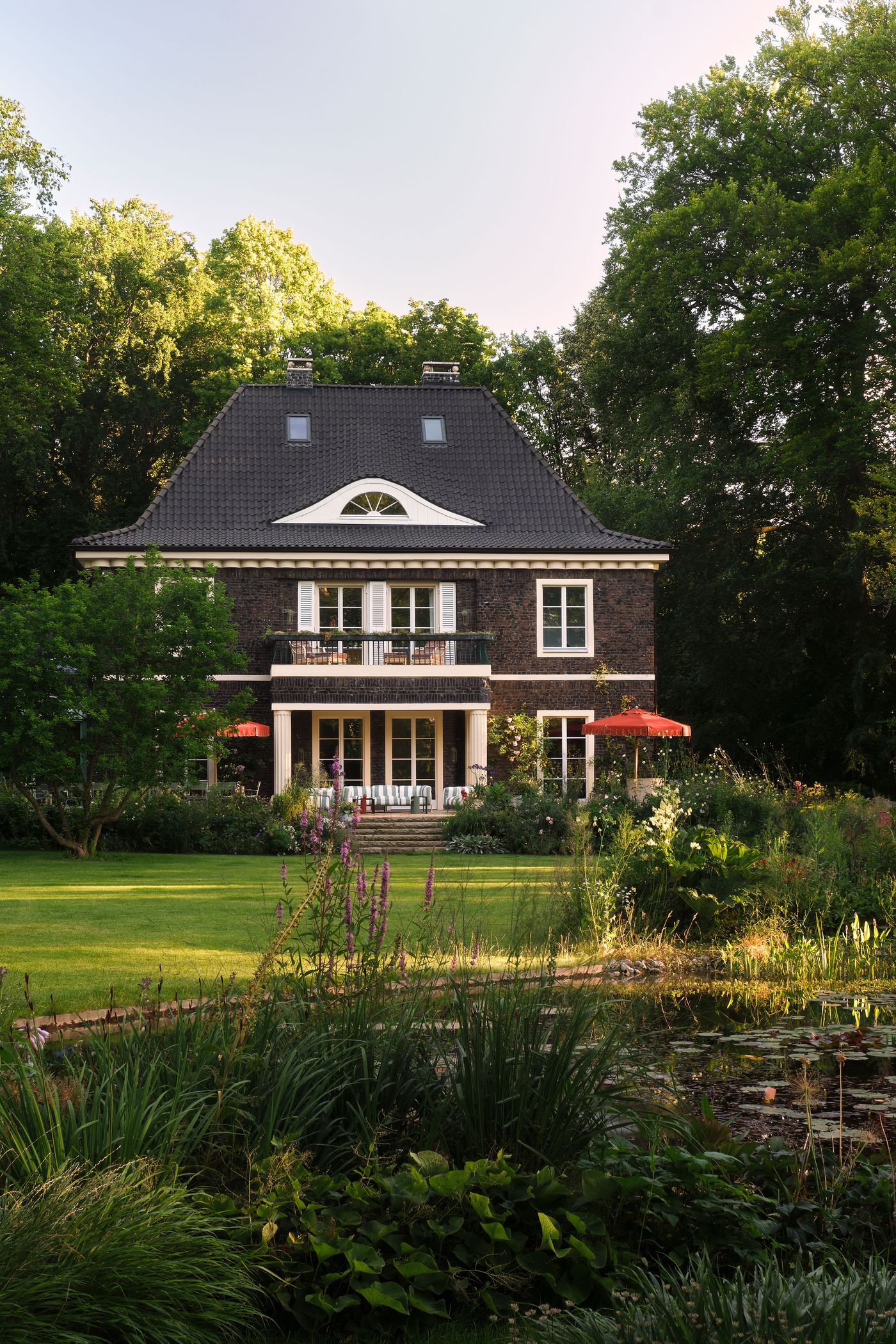 A charming two-story house with a dark tiled roof, surrounded by lush greenery and a vibrant garden, featuring a pond and patio seating. A charming two-story house with a dark tiled roof, surrounded by lush greenery and a vibrant garden, featuring a pond and patio seating.