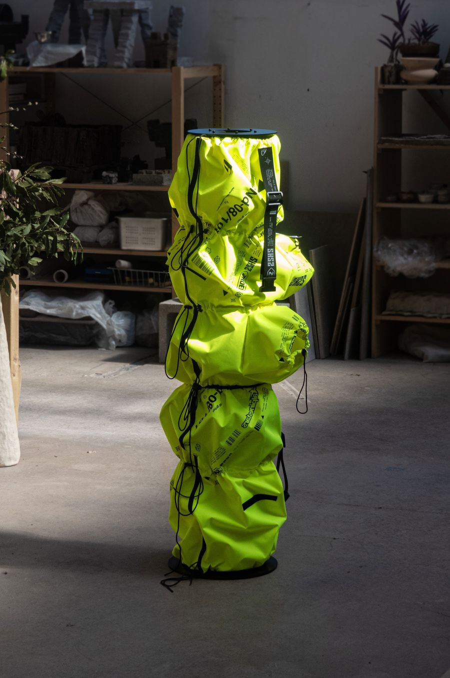 A tall, bright yellow bag with straps and a drawstring, standing in a workshop with shelves of supplies in the background. A tall, bright yellow bag with straps and a drawstring, standing in a workshop with shelves of supplies in the background.