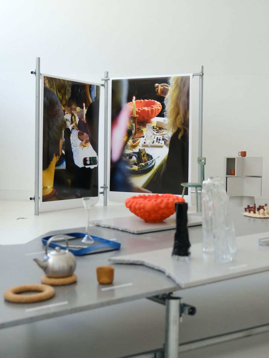 A close-up of the "Sobre Mesa" exhibition by Arquivo Aires Mateus reveals intricate details of the display. On a sleek, light-grey table, unique ceramic pieces and glassware are artfully arranged, including a vibrant orange textured bowl. Large-scale photographic prints in the background depict convivial dining moments, linking the functional design with social interaction. A close-up of the "Sobre Mesa" exhibition by Arquivo Aires Mateus reveals intricate details of the display. On a sleek, light-grey table, unique ceramic pieces and glassware are artfully arranged, including a vibrant orange textured bowl. Large-scale photographic prints in the background depict convivial dining moments, linking the functional design with social interaction.