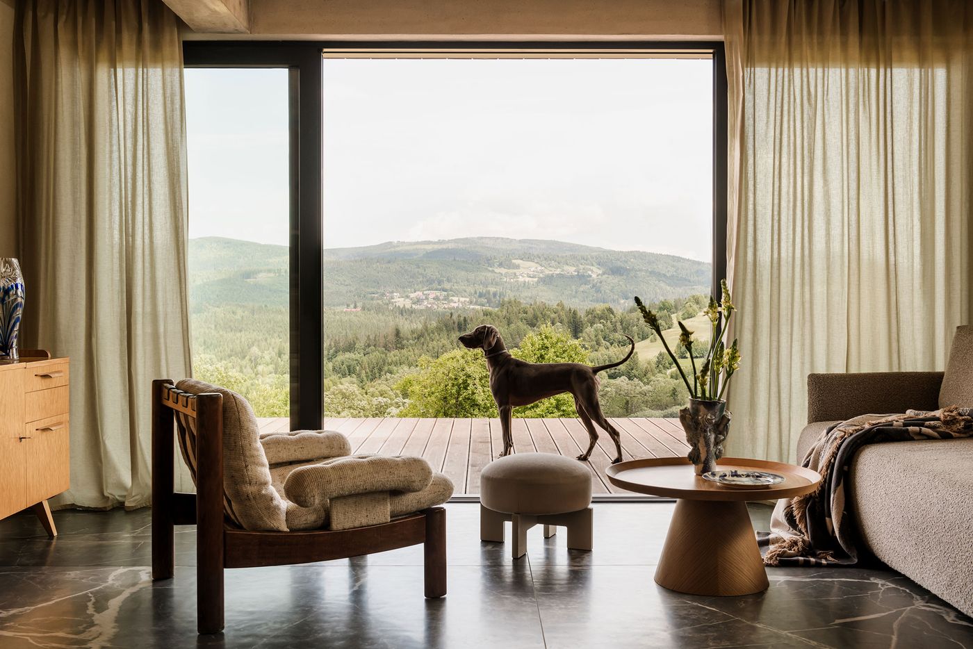A dog stands behind a cushioned stool, gazing through a large window showcasing a lush, green mountainous landscape. A dog stands behind a cushioned stool, gazing through a large window showcasing a lush, green mountainous landscape.