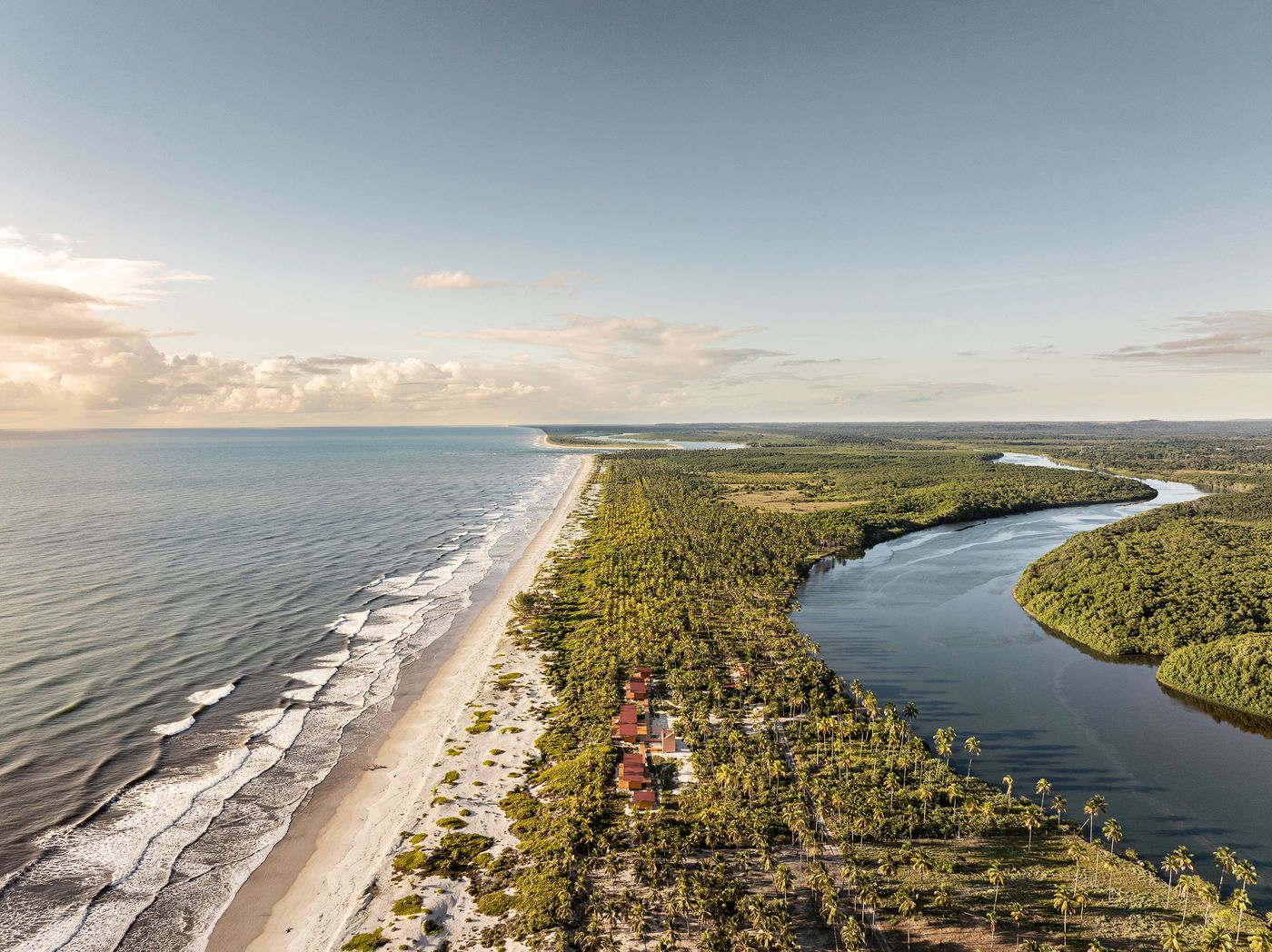 An aerial view showcases a pristine coastline with a curving river meeting the ocean, flanked by dense tropical vegetation and a line of subtle, modular structures. The expansive natural landscape and the discreet architectural intervention suggest a remote, harmonious escape. An aerial view showcases a pristine coastline with a curving river meeting the ocean, flanked by dense tropical vegetation and a line of subtle, modular structures. The expansive natural landscape and the discreet architectural intervention suggest a remote, harmonious escape.