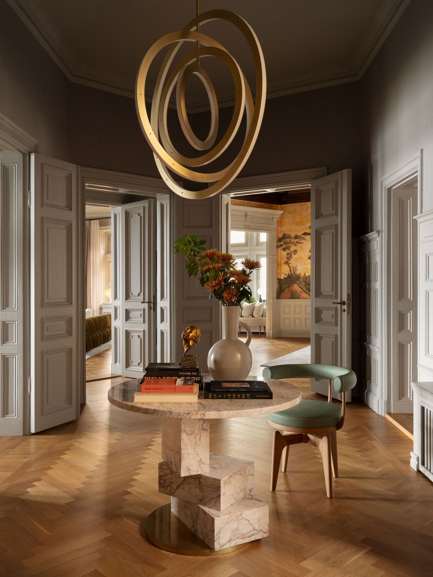 A stylish entryway features a circular gold chandelier, marble table with books, flowers, and a green chair, framed by elegant doors. A stylish entryway features a circular gold chandelier, marble table with books, flowers, and a green chair, framed by elegant doors.