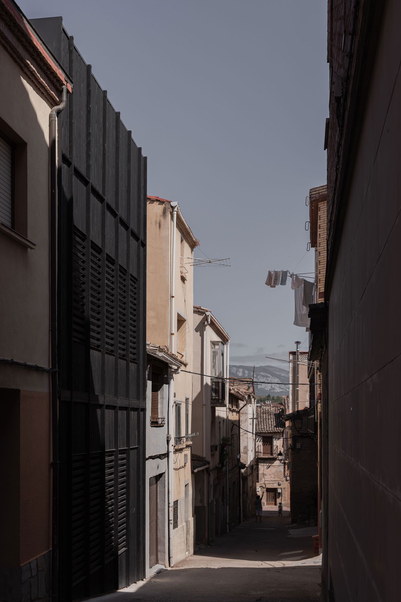 A narrow alleyway lined with contrasting buildings, one modern black facade and vintage structures, laundry hangs overhead against a gray sky. A narrow alleyway lined with contrasting buildings, one modern black facade and vintage structures, laundry hangs overhead against a gray sky.