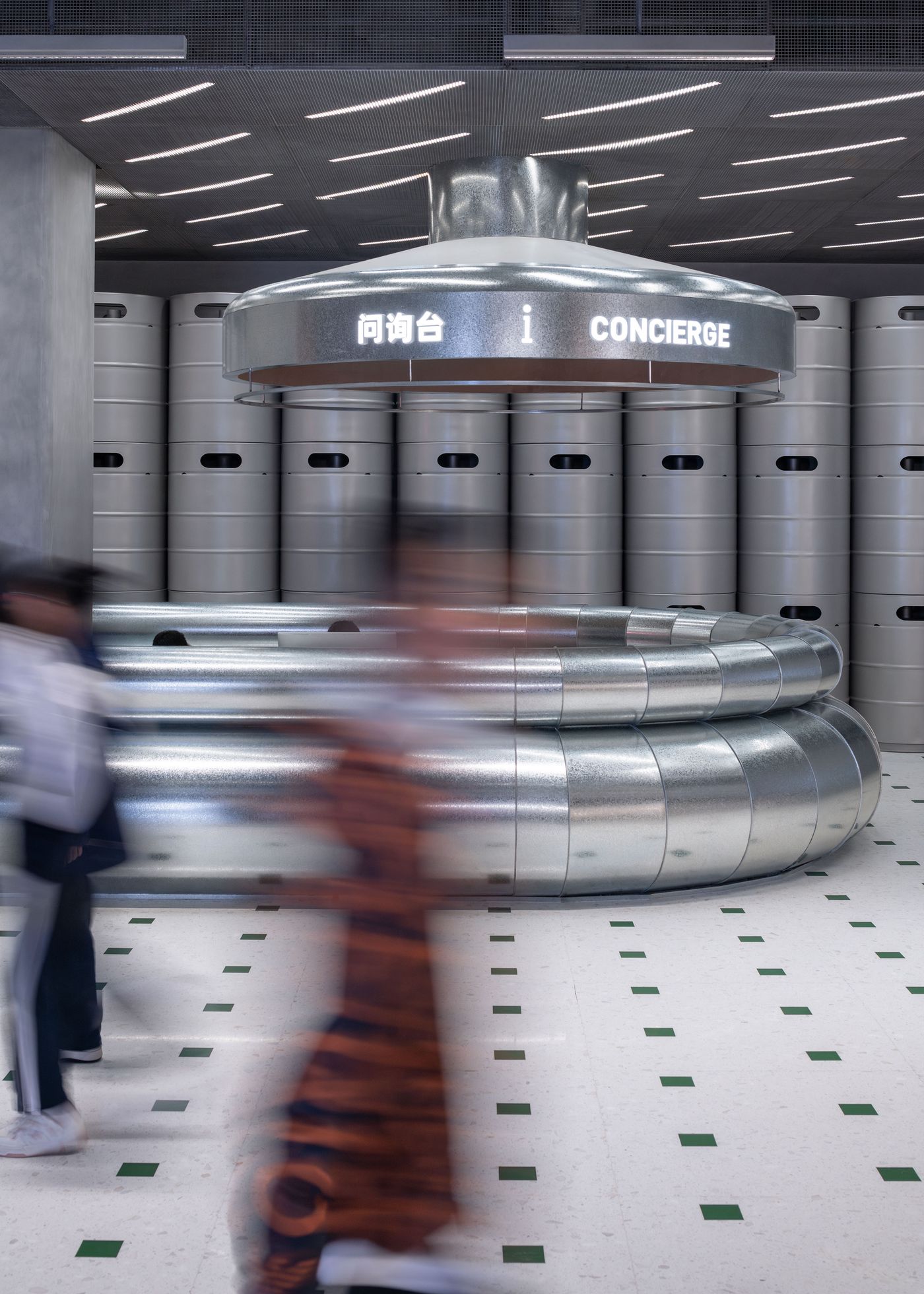 A futuristic concierge desk with metallic design, surrounded by silvery cylindrical structures, and blurred figures walking past.