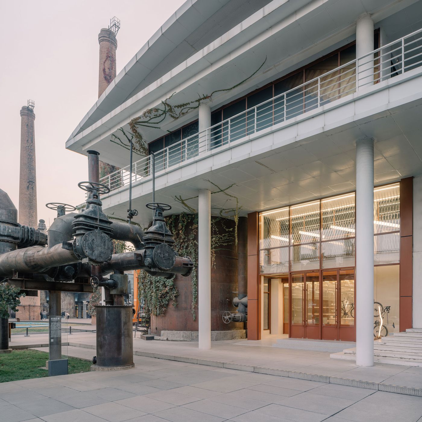 The exterior showcases a brutalist influence with its stark concrete overhangs and prominent white pillars, softened by cascading green ivy that embraces the industrial pipes and oxidized metal structures. The large glass entrance invites light into the space, subtly revealing bicycles within, amidst a raw, industrial park landscape. The exterior showcases a brutalist influence with its stark concrete overhangs and prominent white pillars, softened by cascading green ivy that embraces the industrial pipes and oxidized metal structures. The large glass entrance invites light into the space, subtly revealing bicycles within, amidst a raw, industrial park landscape.