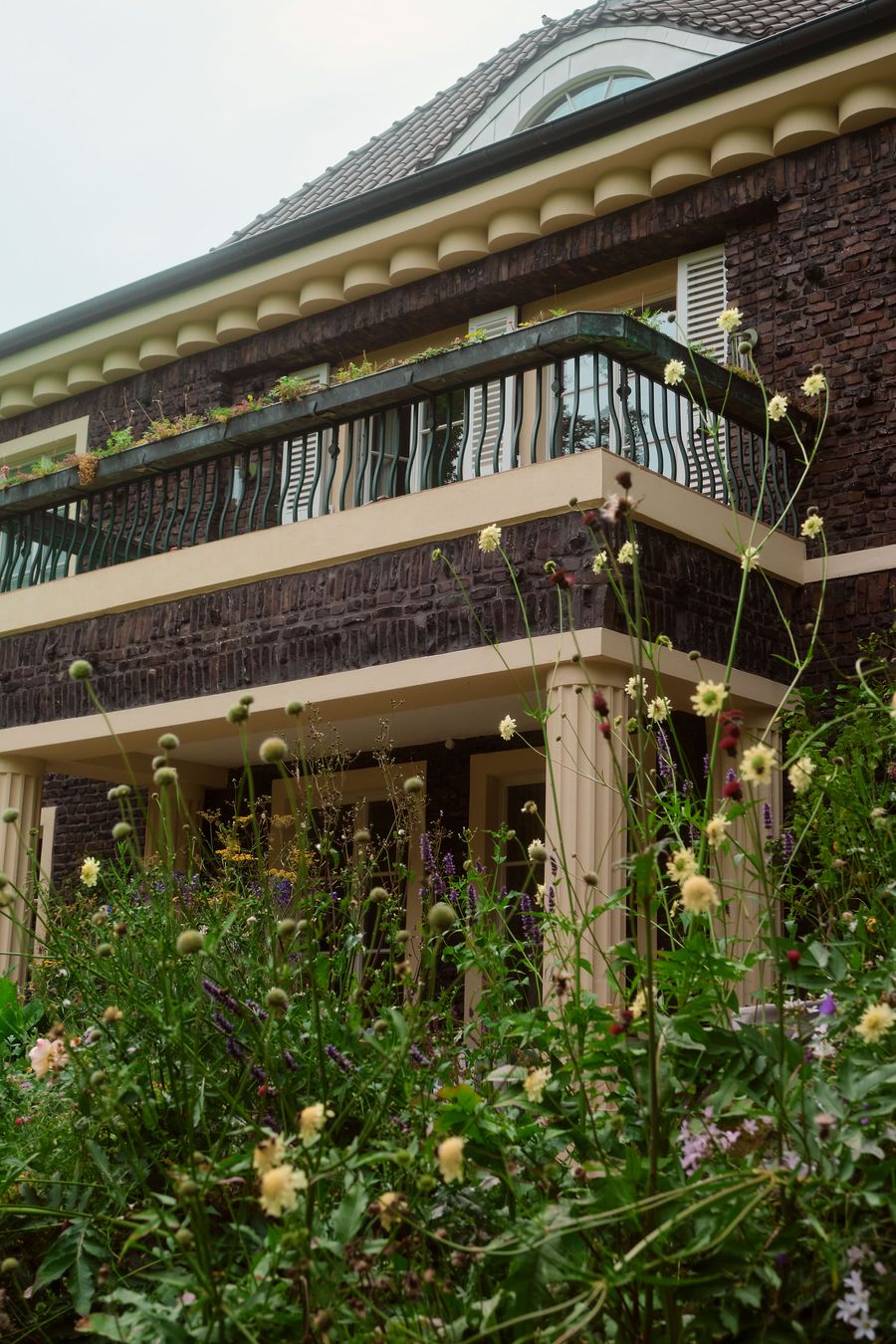 A charming house features a decorative balcony adorned with plants, surrounded by lush flowers and greenery at its base. A charming house features a decorative balcony adorned with plants, surrounded by lush flowers and greenery at its base.