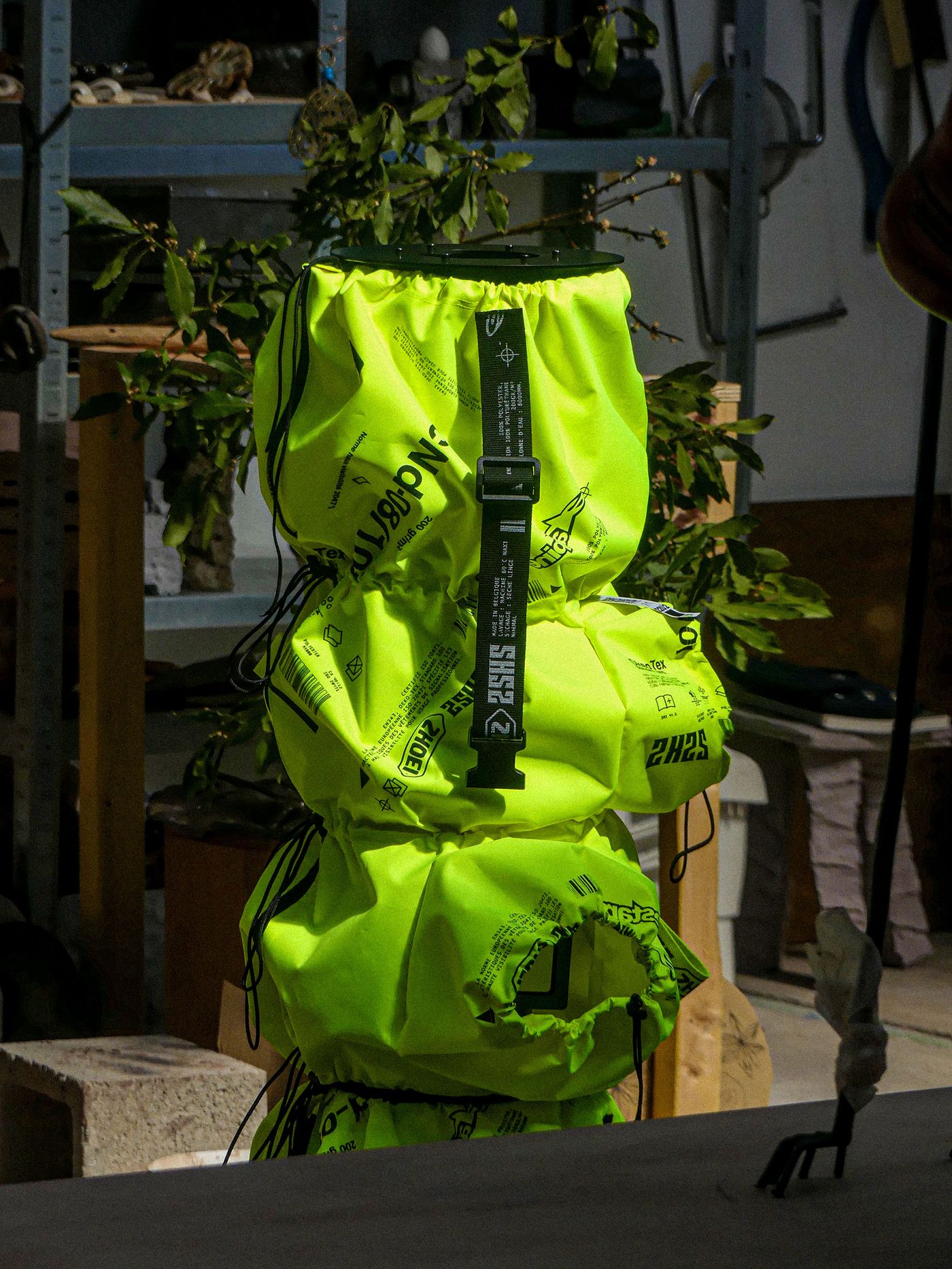 A bright neon yellow bag with black straps and printed designs stands on a table, surrounded by plants and workshop materials.