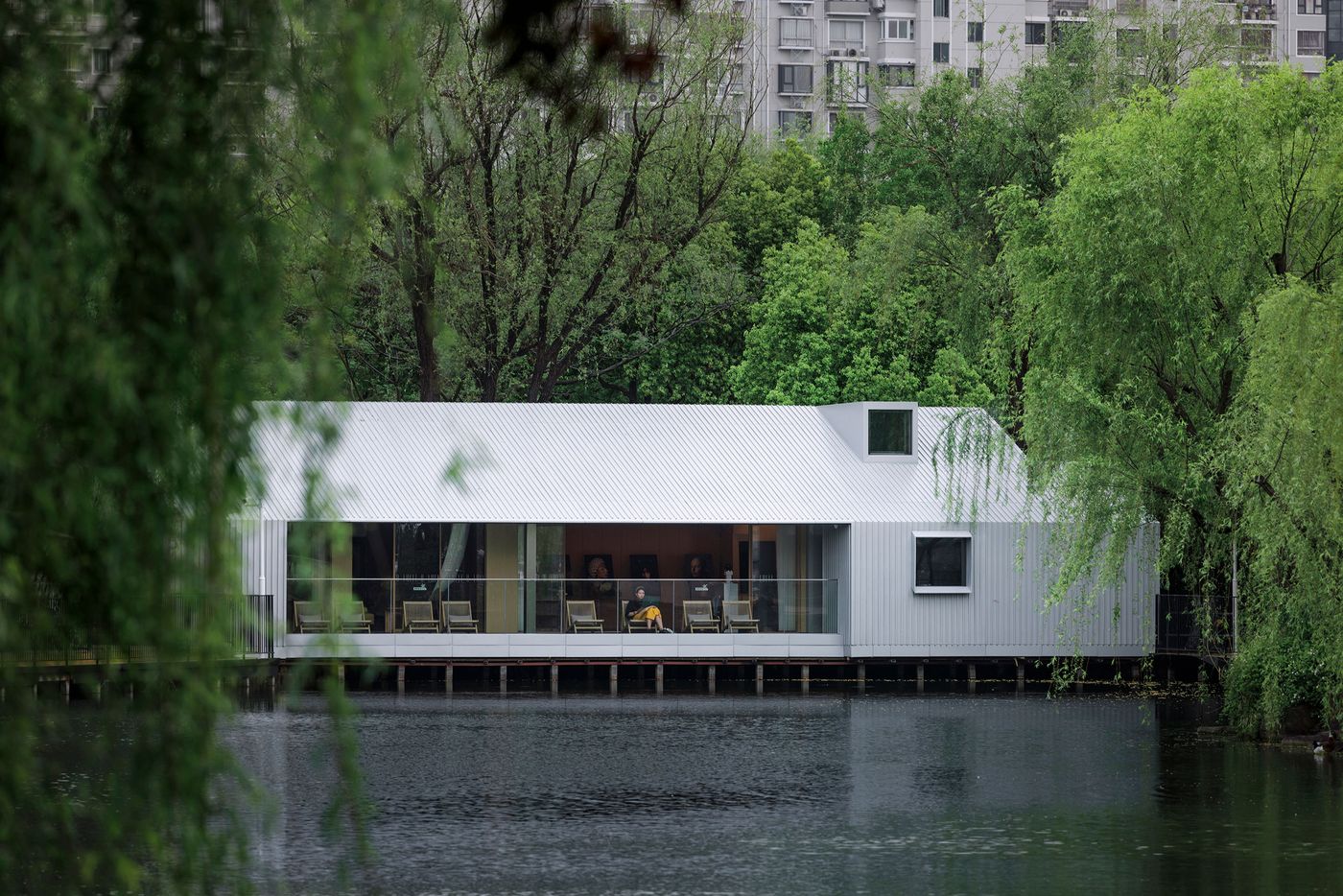 A striking contemporary building by Wutopia Lab, the Lake House in Shanghai, appears to float serenely on dark water. Its minimalist, gabled form is clad in corrugated silver metal, reflecting the overcast sky and lush surrounding foliage. Expansive floor-to-ceiling glass windows reveal a warm, inviting interior with muted furniture and subtle lighting, offering a tranquil sanctuary amidst nature. A striking contemporary building by Wutopia Lab, the Lake House in Shanghai, appears to float serenely on dark water. Its minimalist, gabled form is clad in corrugated silver metal, reflecting the overcast sky and lush surrounding foliage. Expansive floor-to-ceiling glass windows reveal a warm, inviting interior with muted furniture and subtle lighting, offering a tranquil sanctuary amidst nature.