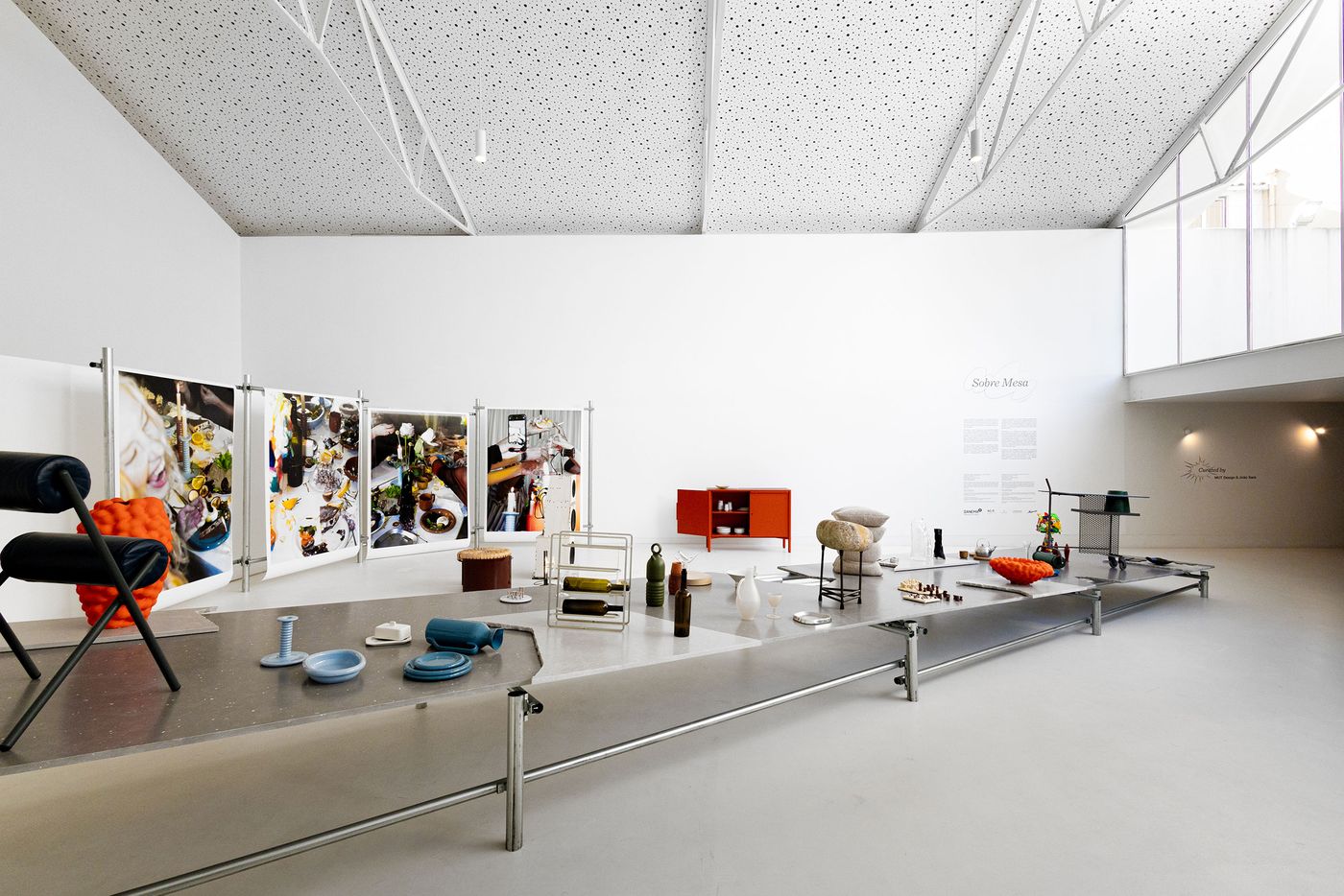 The "Sobre Mesa" exhibition by Arquivo Aires Mateus features a long, low display table showcasing a diverse collection of contemporary tableware and ceramic objects in muted and vibrant tones. Behind it, large photographic panels depict lively dining scenes, creating a dynamic interplay between the exhibited objects and their potential use, set within a spacious, modern gallery. The "Sobre Mesa" exhibition by Arquivo Aires Mateus features a long, low display table showcasing a diverse collection of contemporary tableware and ceramic objects in muted and vibrant tones. Behind it, large photographic panels depict lively dining scenes, creating a dynamic interplay between the exhibited objects and their potential use, set within a spacious, modern gallery.