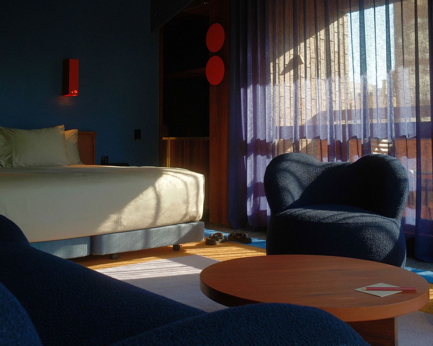 A cozy hotel room featuring a bed, blue armchairs, wooden table, and soft lighting from sheer curtains, creating a warm atmosphere. A cozy hotel room featuring a bed, blue armchairs, wooden table, and soft lighting from sheer curtains, creating a warm atmosphere.