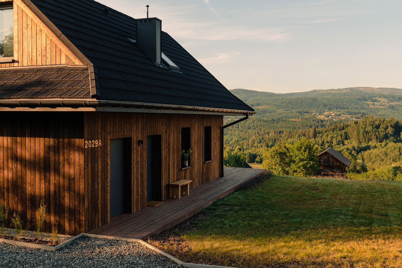 A modern wooden house with a dark roof set against a green valley, featuring a wooden deck and scenic mountainous backdrop. A modern wooden house with a dark roof set against a green valley, featuring a wooden deck and scenic mountainous backdrop.