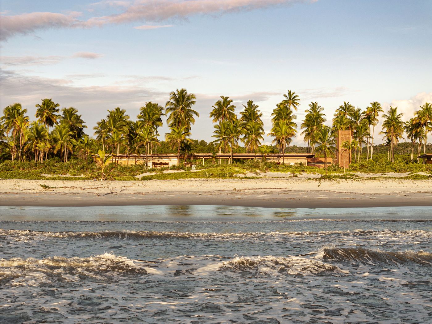 From the ocean, a low-profile resort blends into the lush coastal landscape, nestled behind a sandy beach and a line of palm trees. The gentle waves in the foreground contribute to the tranquil, authentic seaside atmosphere, prioritizing harmony with nature. From the ocean, a low-profile resort blends into the lush coastal landscape, nestled behind a sandy beach and a line of palm trees. The gentle waves in the foreground contribute to the tranquil, authentic seaside atmosphere, prioritizing harmony with nature.