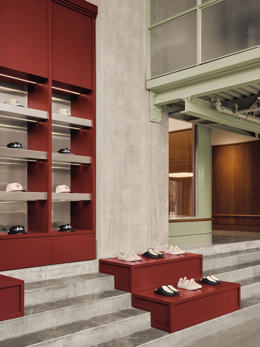 A modern retail space featuring red display shelves with hats and sneakers arranged on steps, against a concrete wall and green accents. A modern retail space featuring red display shelves with hats and sneakers arranged on steps, against a concrete wall and green accents.