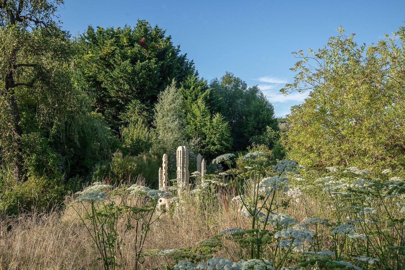A serene garden scene featuring tall, green trees and white flowering plants surrounding unique stone sculptures under a clear blue sky. A serene garden scene featuring tall, green trees and white flowering plants surrounding unique stone sculptures under a clear blue sky.