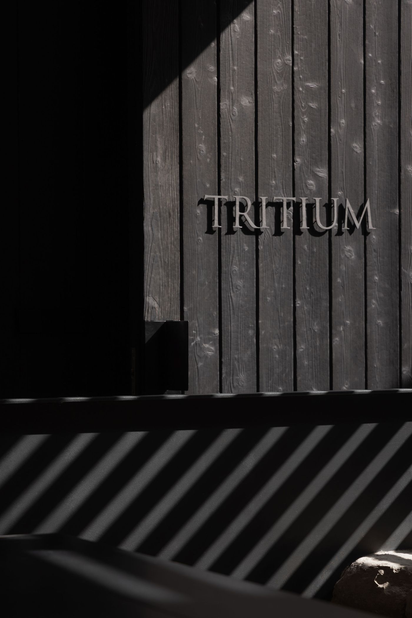 Dark wooden wall with the word "TRITIUM" carved in, contrasted by diagonal light and shadow patterns across the foreground. Dark wooden wall with the word "TRITIUM" carved in, contrasted by diagonal light and shadow patterns across the foreground.