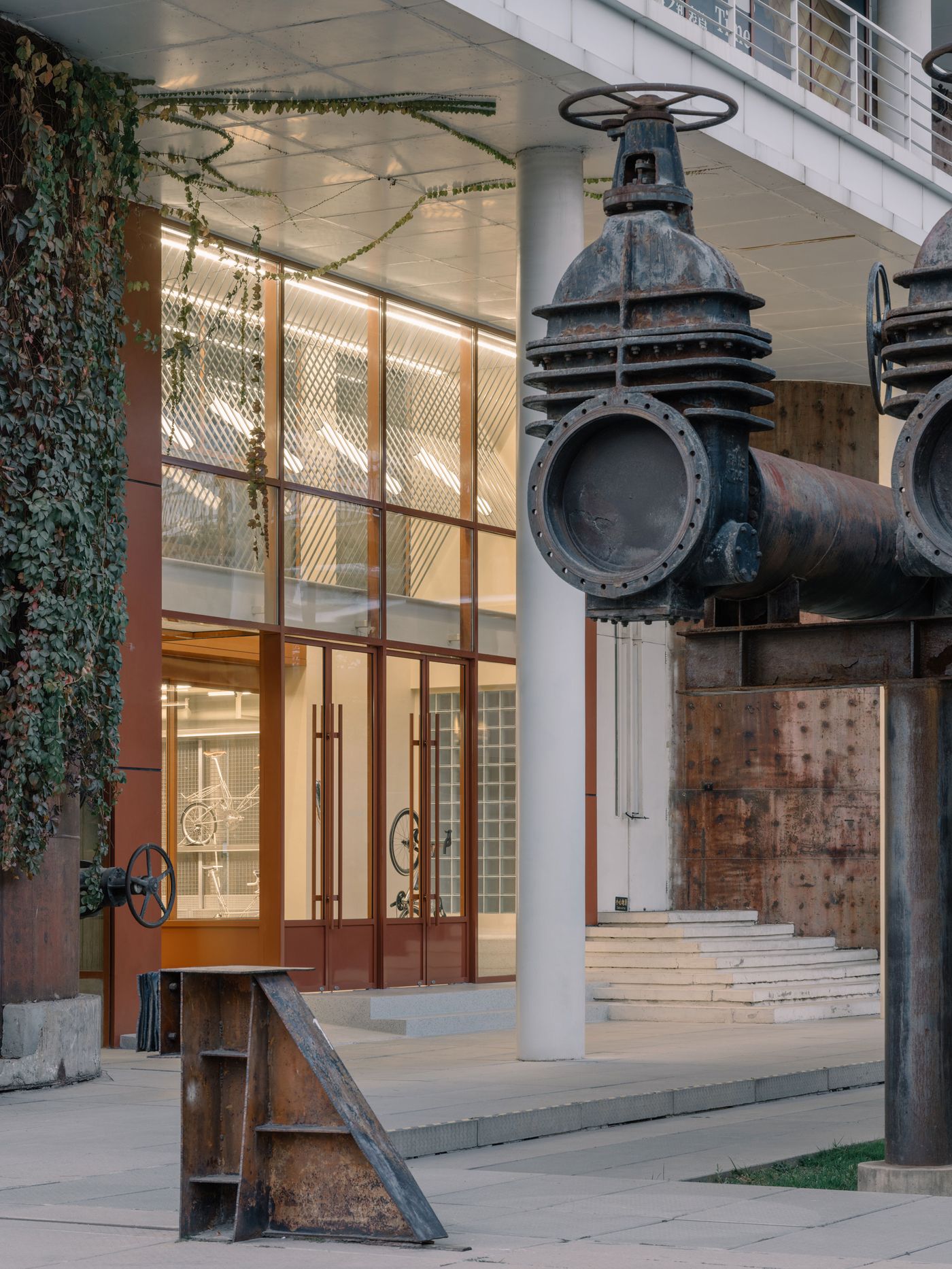 A close-up of the entrance highlights the dramatic scale of repurposed industrial elements, including colossal rusted valves and pipes, juxtaposed against the building's sleek, modern glass facade. The interplay of the raw, textured metal and the smooth, transparent glass, alongside the creeping ivy, embodies a compelling industrial-biophilic design. A close-up of the entrance highlights the dramatic scale of repurposed industrial elements, including colossal rusted valves and pipes, juxtaposed against the building's sleek, modern glass facade. The interplay of the raw, textured metal and the smooth, transparent glass, alongside the creeping ivy, embodies a compelling industrial-biophilic design.