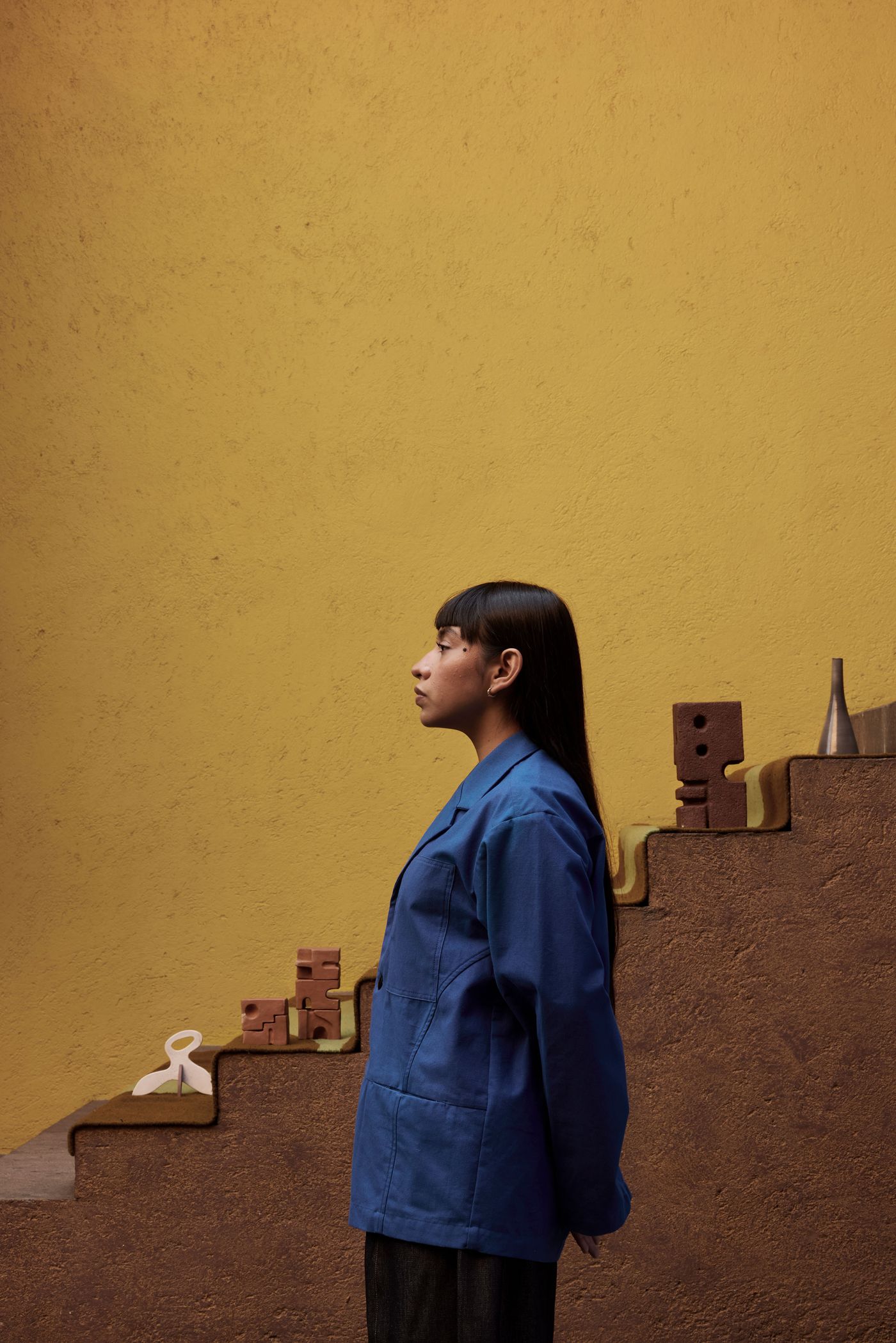 A person in a blue coat stands beside colorful abstract objects on stylized steps against a warm yellow wall.