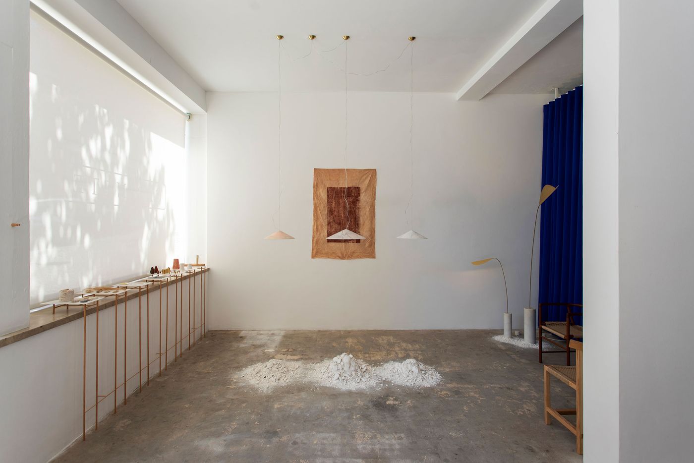 An industrial-minimalist gallery space by Bamansure features a stark white backdrop and concrete floor. Three piles of white material on the floor form a sculptural focal point, complemented by a hanging abstract artwork in earthy tones. Delicate pendant lights and a deep blue curtain add splashes of color, creating a contemplative atmosphere focused on raw materials and form. An industrial-minimalist gallery space by Bamansure features a stark white backdrop and concrete floor. Three piles of white material on the floor form a sculptural focal point, complemented by a hanging abstract artwork in earthy tones. Delicate pendant lights and a deep blue curtain add splashes of color, creating a contemplative atmosphere focused on raw materials and form.
