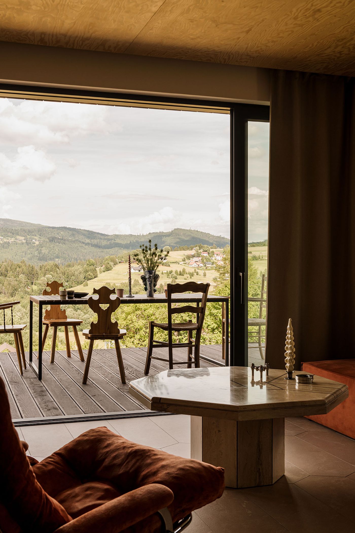 Stylish interior with a cozy orange chair, a marble table, and a scenic view of rolling hills and trees through large glass doors. Stylish interior with a cozy orange chair, a marble table, and a scenic view of rolling hills and trees through large glass doors.