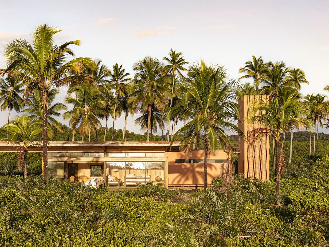 A single-storey modern dwelling, partially concealed by lush tropical foliage, features expansive glass walls and warm, earthy-toned cladding, including a distinctive brick-like tower element. The structure's horizontal emphasis and material palette create a dialogue with the vibrant natural surroundings, characteristic of biophilic design. A single-storey modern dwelling, partially concealed by lush tropical foliage, features expansive glass walls and warm, earthy-toned cladding, including a distinctive brick-like tower element. The structure's horizontal emphasis and material palette create a dialogue with the vibrant natural surroundings, characteristic of biophilic design.