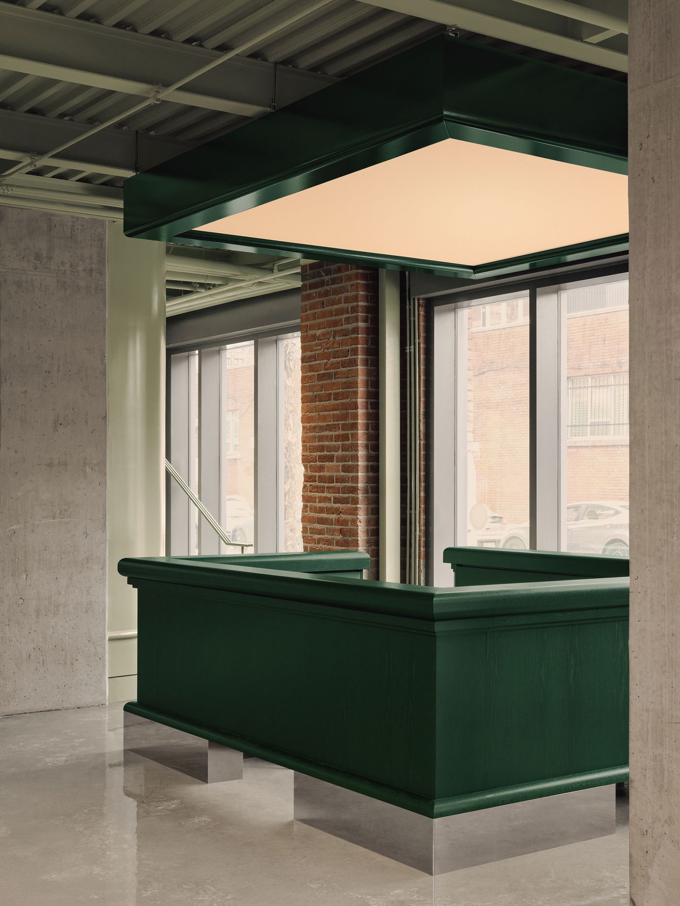 Modern reception area with a green wooden desk, illuminated by a hanging light fixture, featuring exposed brick and large windows.