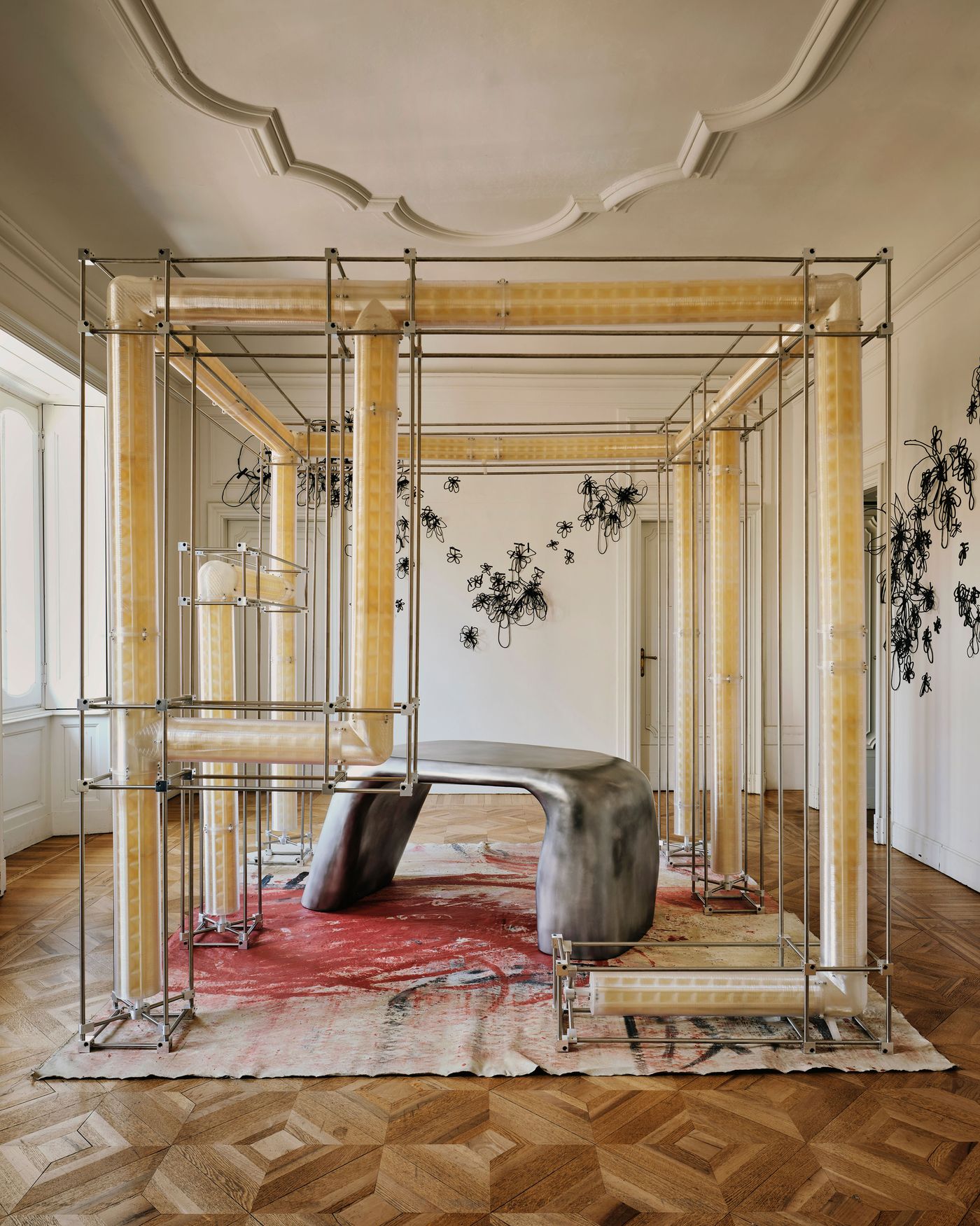 An artistic installation features yellow tubular structures and a sleek, gray table on a patterned rug, set in an elegant room.