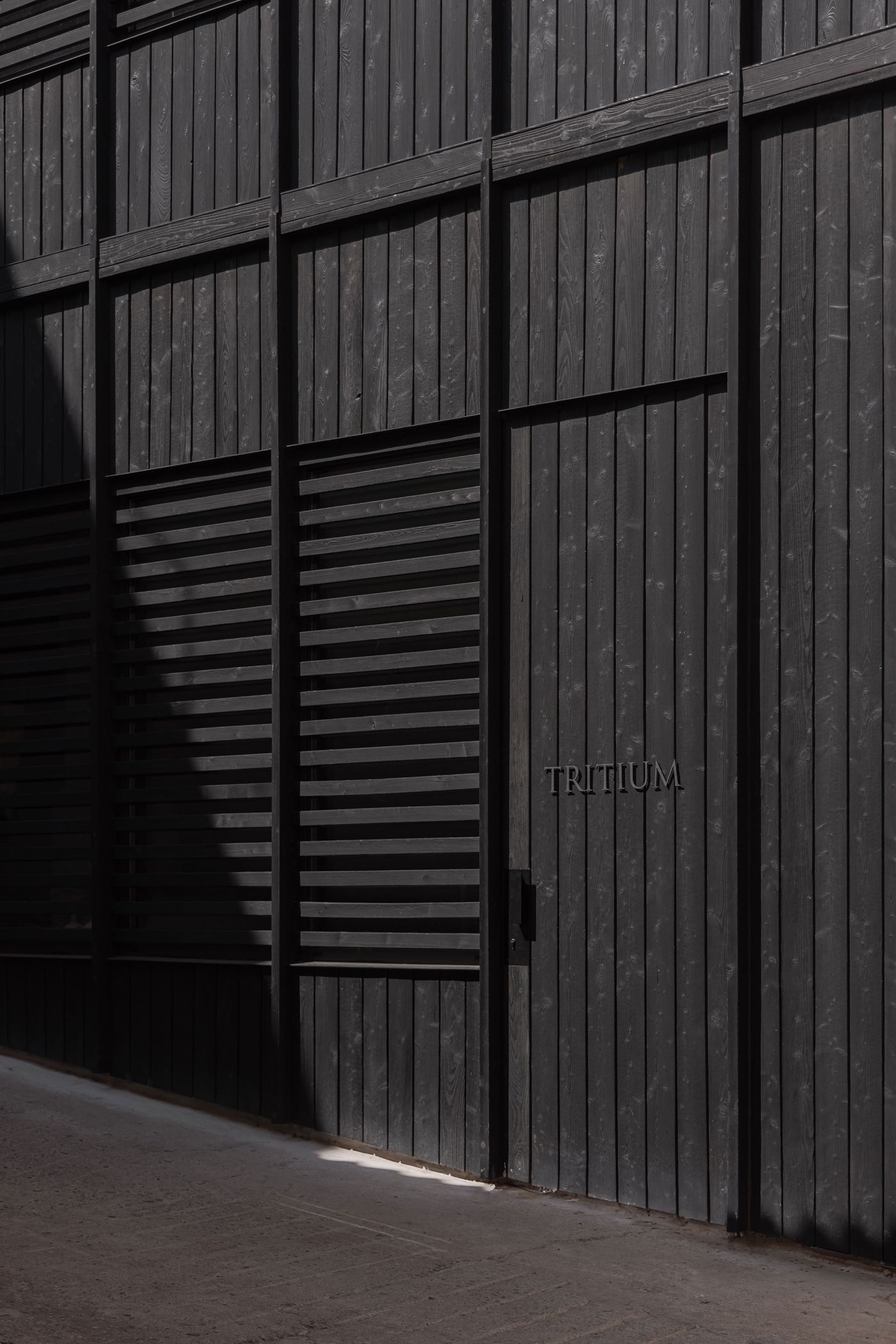 A black wooden wall with a narrow door labeled "TRITIUM," featuring vertical planks and horizontal slats, partially shaded by sunlight. A black wooden wall with a narrow door labeled "TRITIUM," featuring vertical planks and horizontal slats, partially shaded by sunlight.