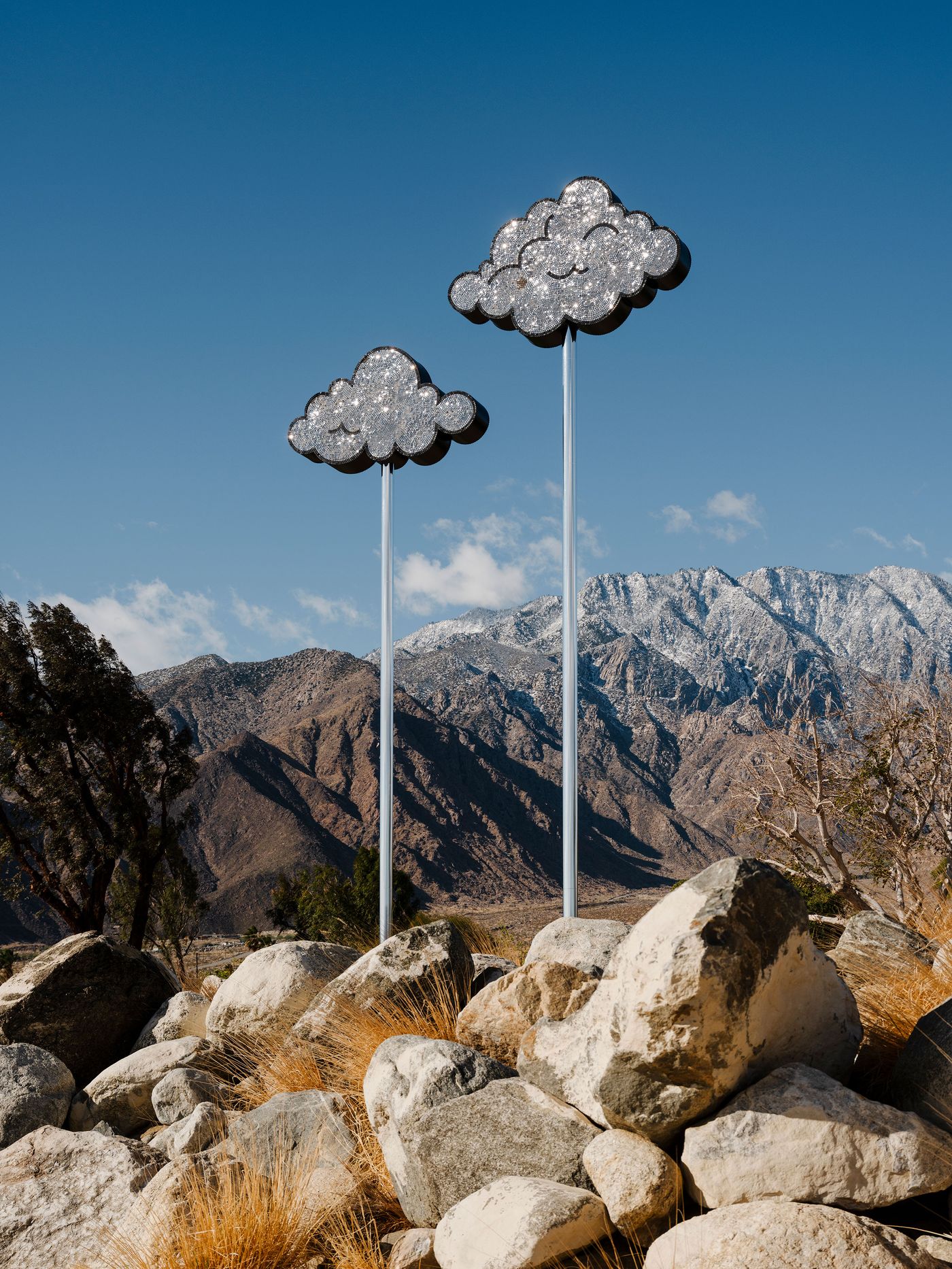 Two glittering cloud sculptures on tall poles stand against a clear blue sky and snow-capped mountains, surrounded by rocky terrain. Two glittering cloud sculptures on tall poles stand against a clear blue sky and snow-capped mountains, surrounded by rocky terrain.