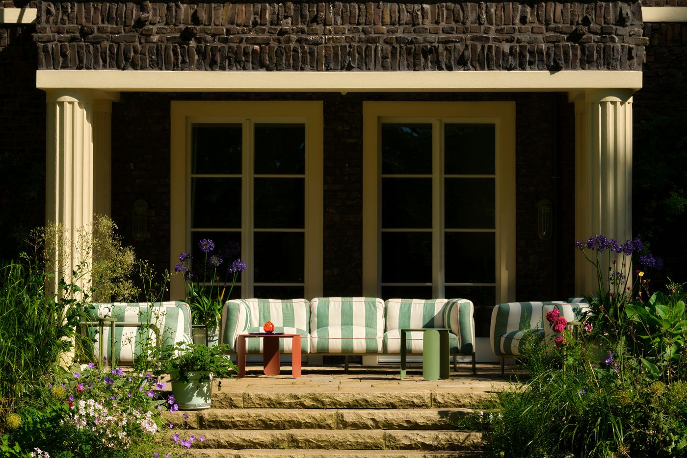 A cozy porch featuring green and white striped sofas, colorful side tables, and vibrant flowers, framed by tall stone columns. A cozy porch featuring green and white striped sofas, colorful side tables, and vibrant flowers, framed by tall stone columns.