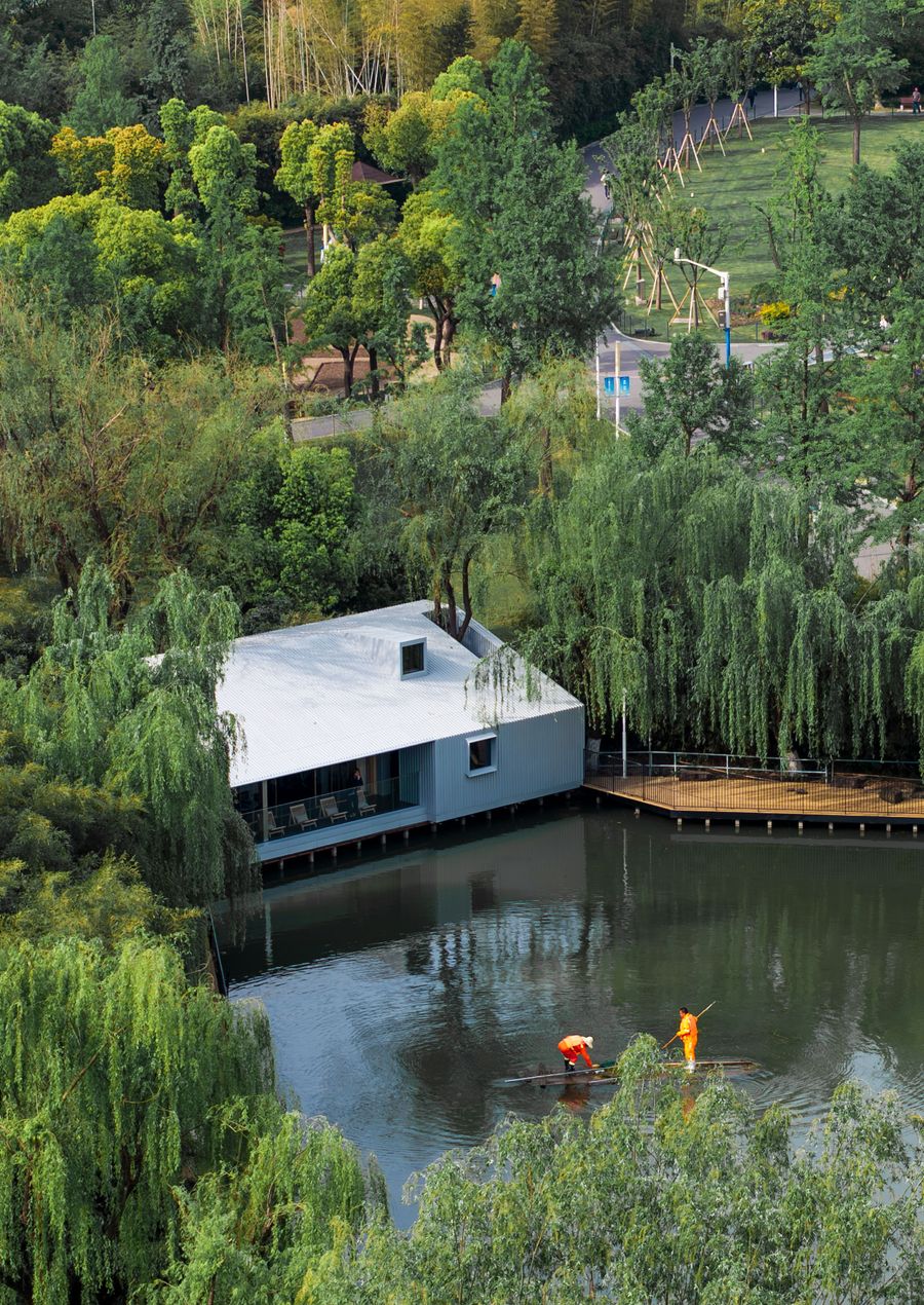 An aerial view showcases Wutopia Lab's Lake House nestled within a vibrant green landscape in Shanghai, its clean lines and white corrugated facade contrasting with the organic forms of weeping willows. The structure extends over the calm lake, with a wooden deck connecting to the shore. Two individuals in orange life vests are visible on the water, highlighting the serene, biophilic integration of the architecture. An aerial view showcases Wutopia Lab's Lake House nestled within a vibrant green landscape in Shanghai, its clean lines and white corrugated facade contrasting with the organic forms of weeping willows. The structure extends over the calm lake, with a wooden deck connecting to the shore. Two individuals in orange life vests are visible on the water, highlighting the serene, biophilic integration of the architecture.