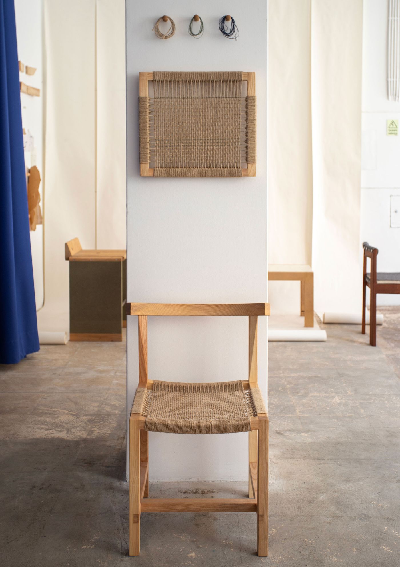 A detail shot from Bamansure's exhibit showcases a natural wood chair with a woven rush seat and back, embodying a rustic-minimalist aesthetic. Behind it, a corresponding square woven panel hangs on a white partition. The simple, honest construction and warm textures highlight a focus on traditional craftsmanship and natural materials within an open, industrial setting.