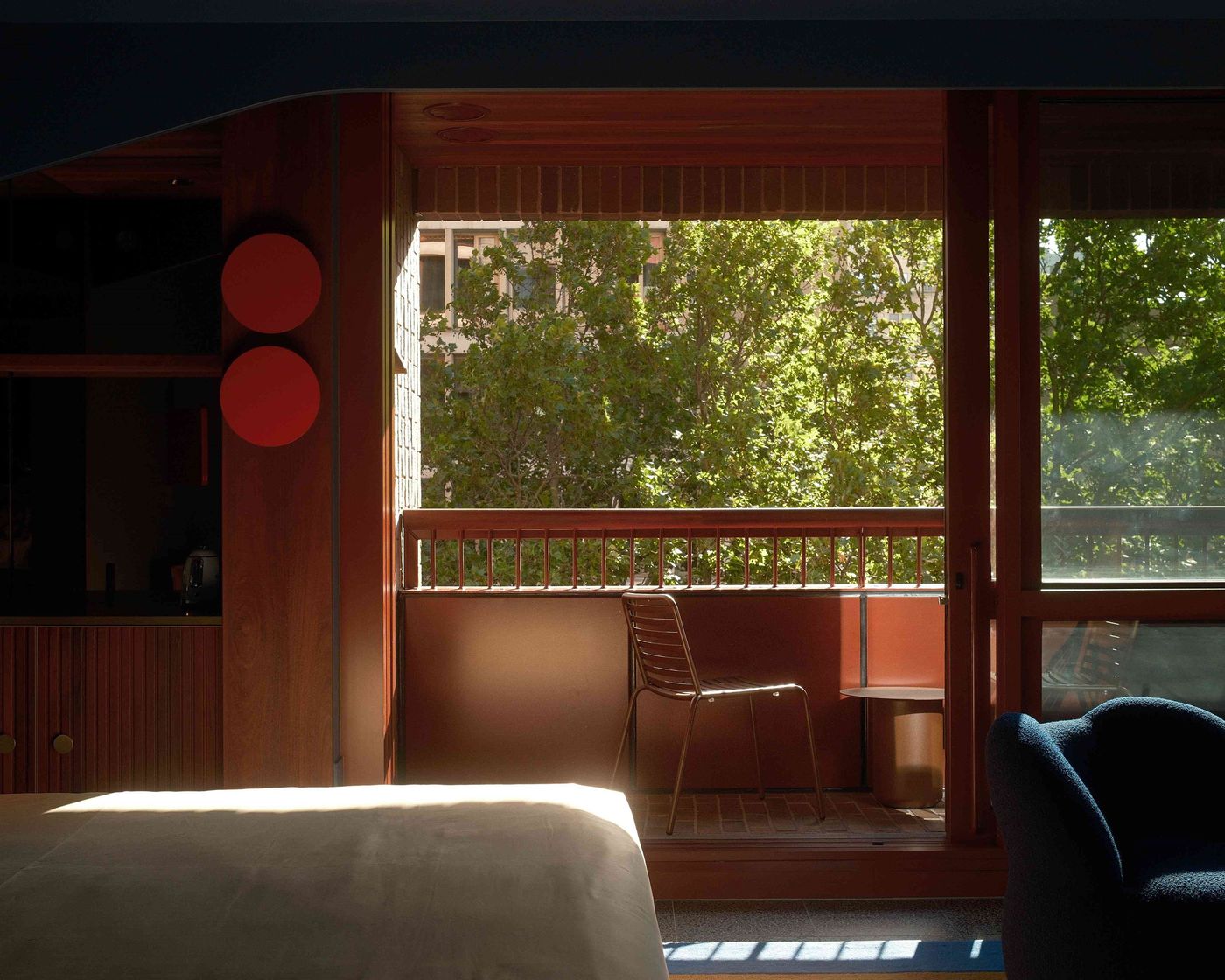 Sunlit hotel room with wooden accents, showcasing a balcony with a chair and table, framed by lush greenery outside.