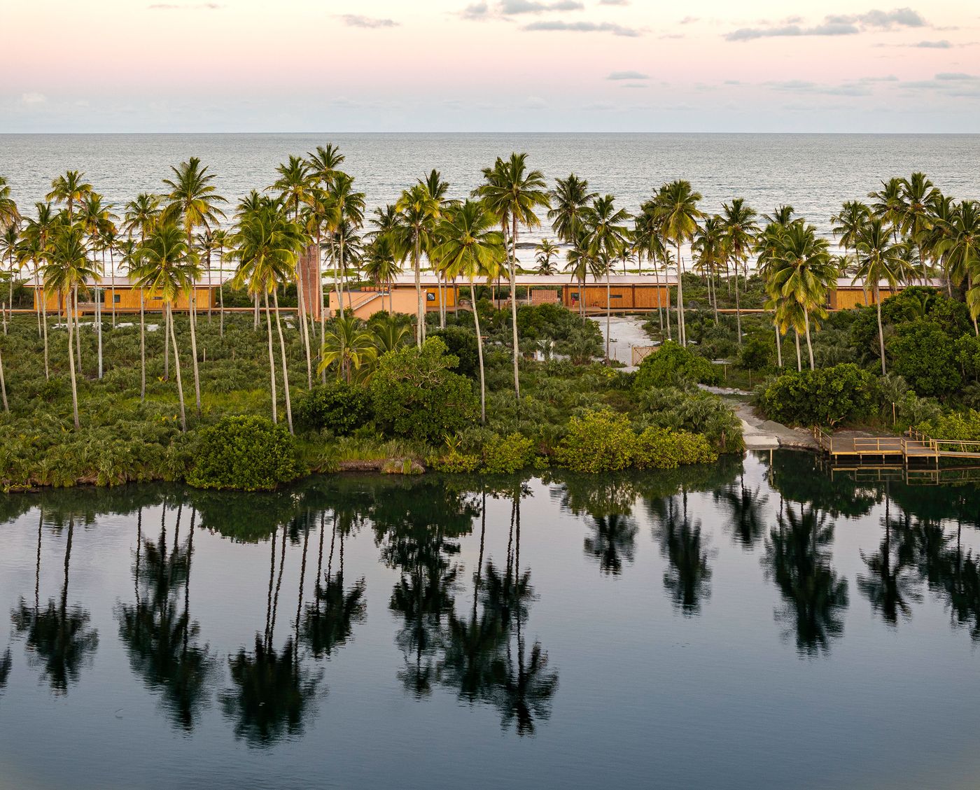 A tropical coastal scene features a tranquil laggon reflecting palm trees and the sky, with low-slung buildings visible amidst lush greenery. The warm wooden tones of the structures blend harmoniously with the natural landscape, evoking a sense of secluded, biophilic luxury. A tropical coastal scene features a tranquil laggon reflecting palm trees and the sky, with low-slung buildings visible amidst lush greenery. The warm wooden tones of the structures blend harmoniously with the natural landscape, evoking a sense of secluded, biophilic luxury.