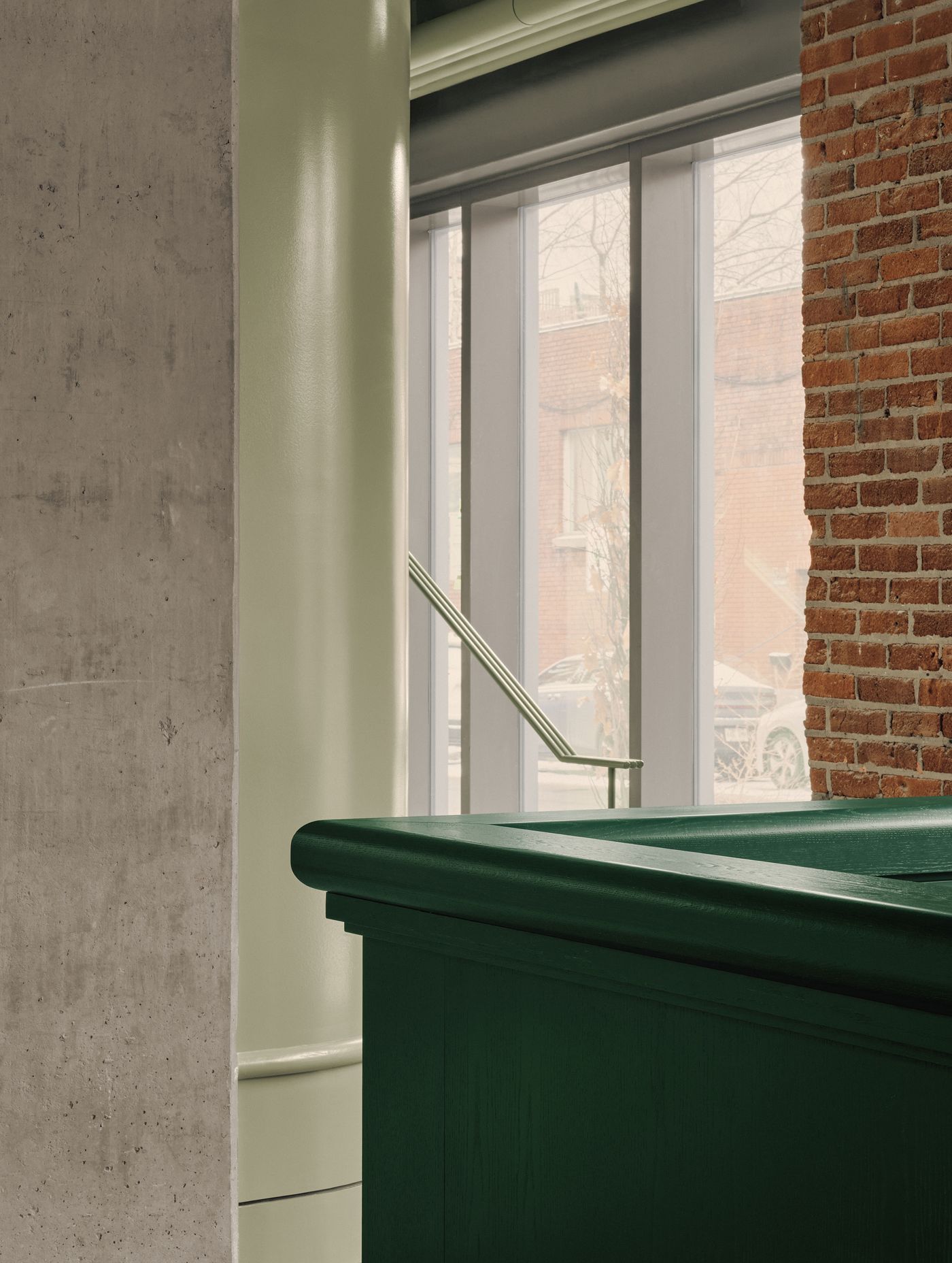 A modern interior featuring a green wooden counter, green column, and exposed red brick wall, with large windows allowing natural light.