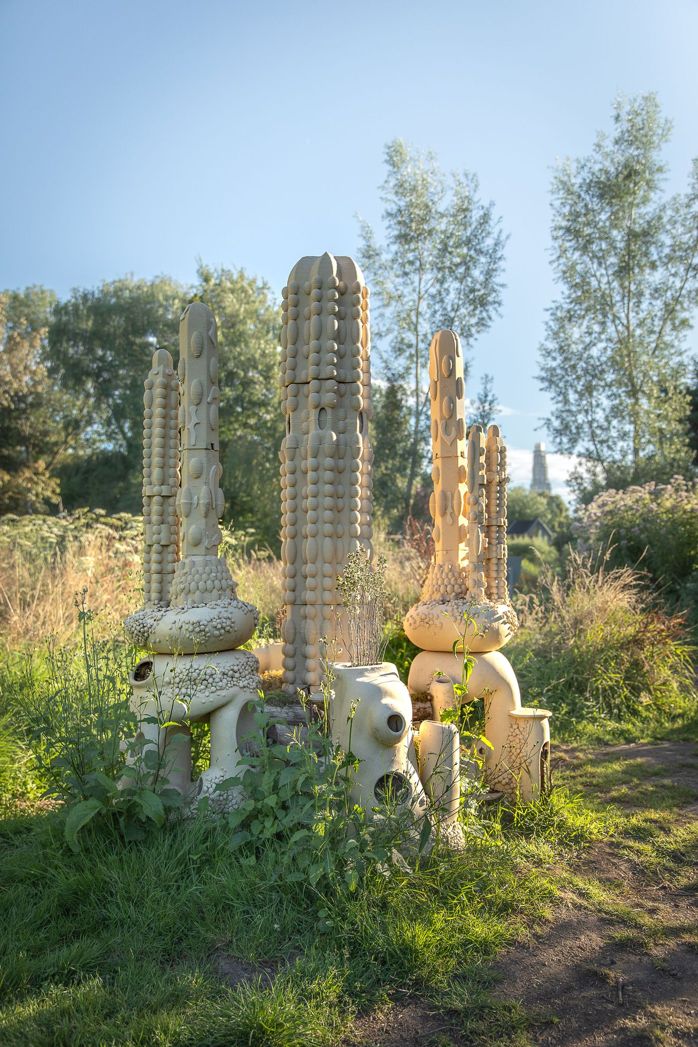 A cluster of beige, abstract sculptures resembling cacti amidst lush greenery under a clear blue sky. A cluster of beige, abstract sculptures resembling cacti amidst lush greenery under a clear blue sky.
