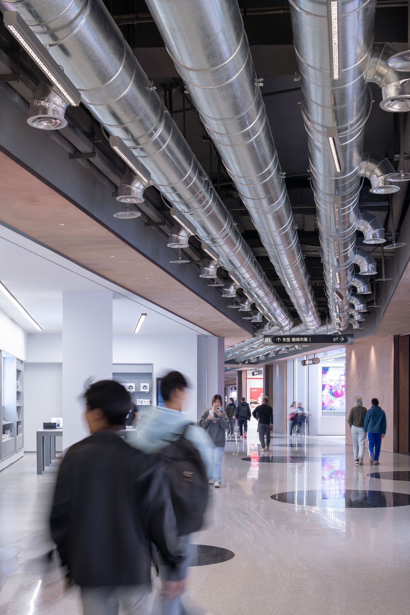A modern interior space featuring polished floors, overhead ductwork, and people walking past contemporary storefronts and displays.