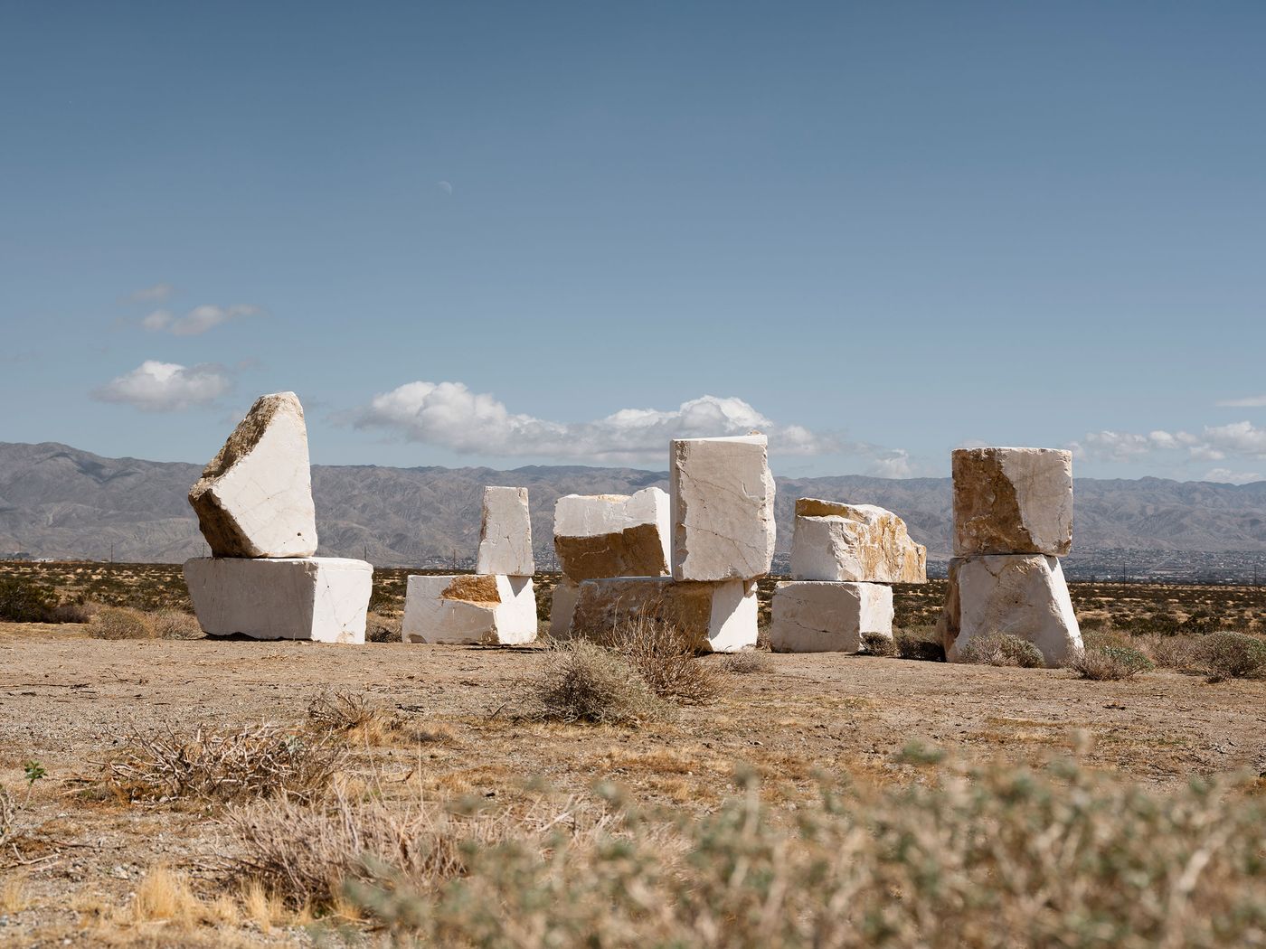 A group of large white stone blocks arranged in an art installation, set against a desert landscape and mountain backdrop under a blue sky. A group of large white stone blocks arranged in an art installation, set against a desert landscape and mountain backdrop under a blue sky.