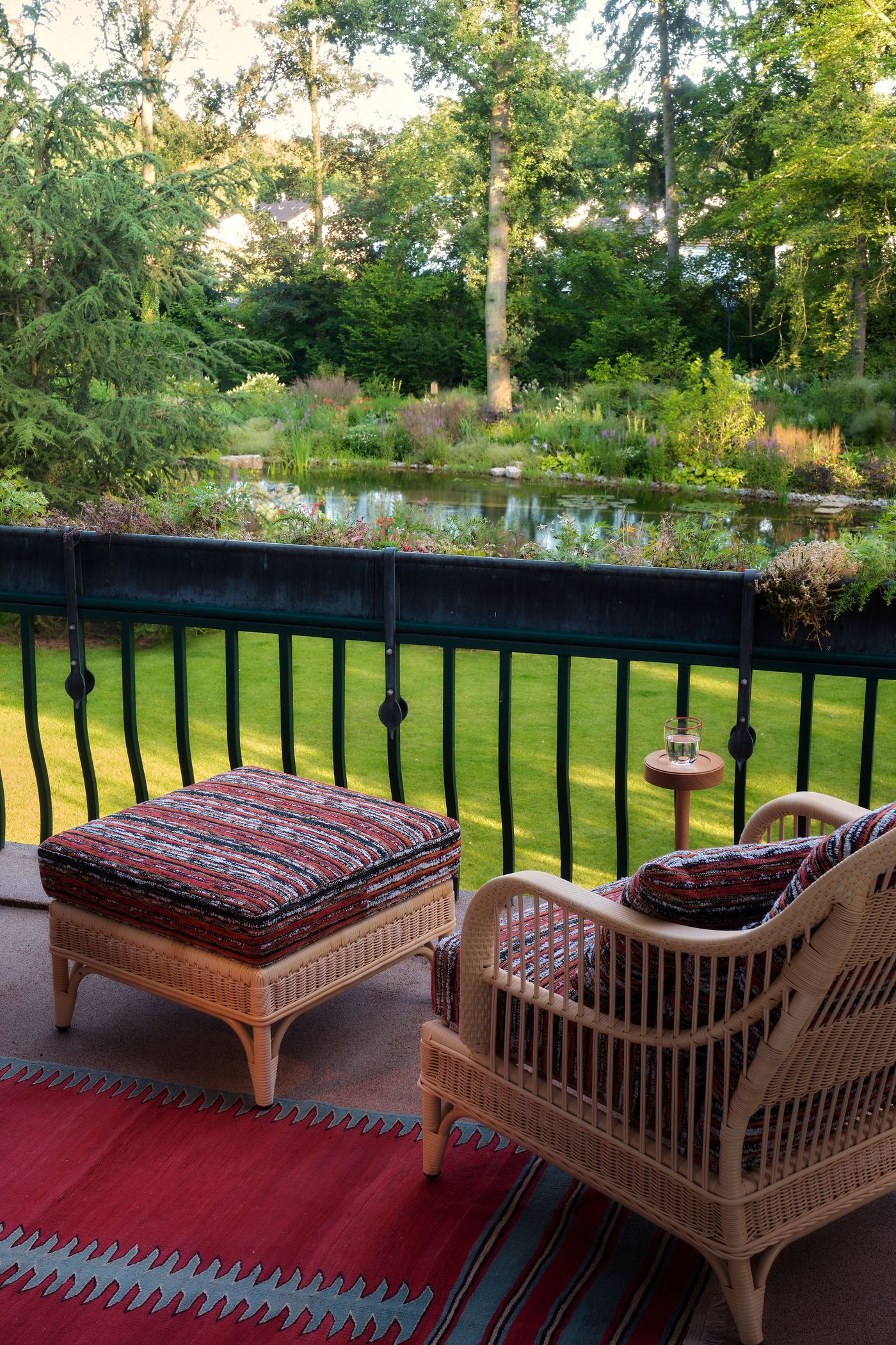 A cozy balcony with wicker seating, colorful cushions, and a small table next to a serene garden pond surrounded by lush greenery.