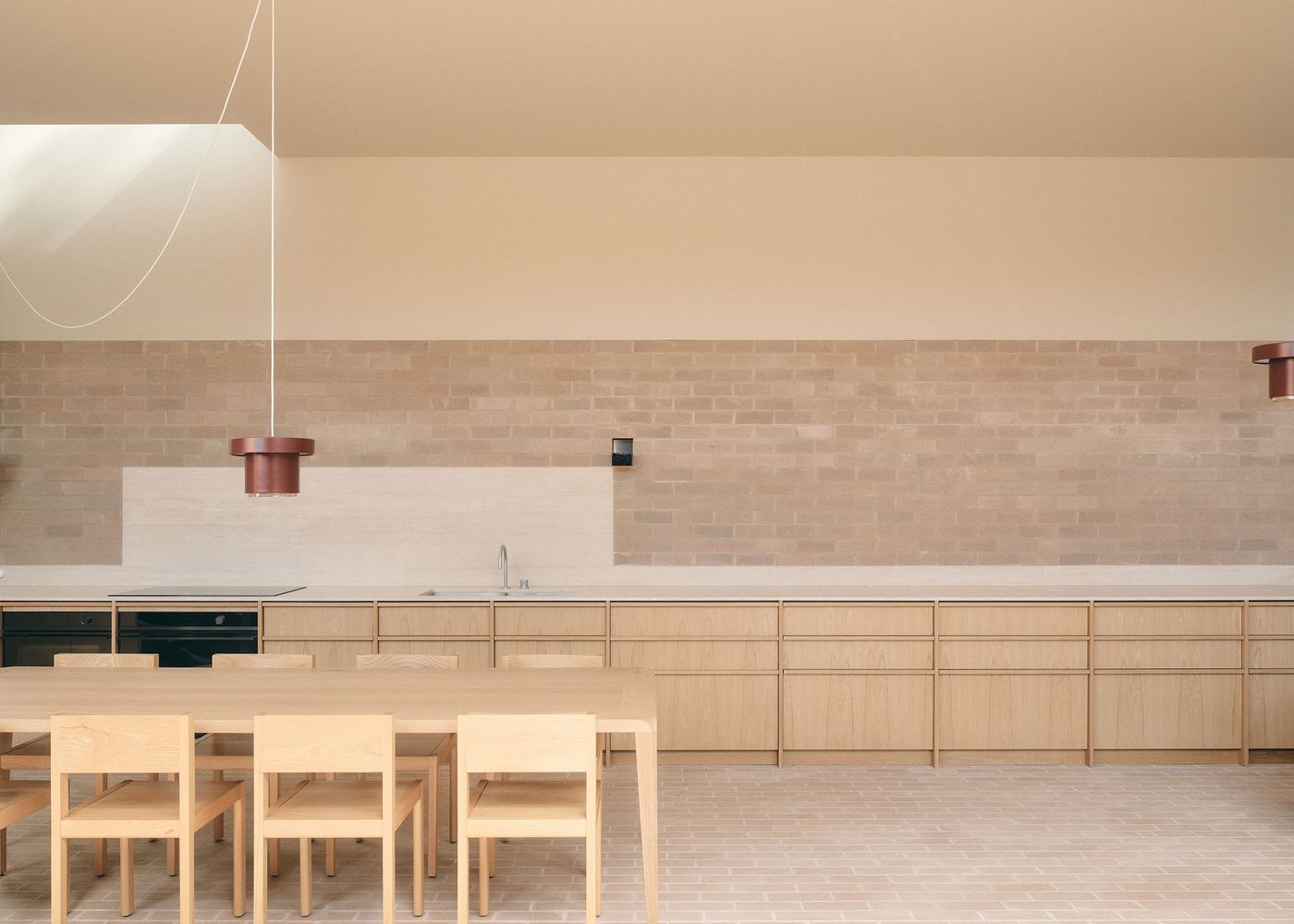 Modern kitchen featuring wooden cabinetry, a long wooden dining table, and minimalist pendant lights against a brick wall. Modern kitchen featuring wooden cabinetry, a long wooden dining table, and minimalist pendant lights against a brick wall.