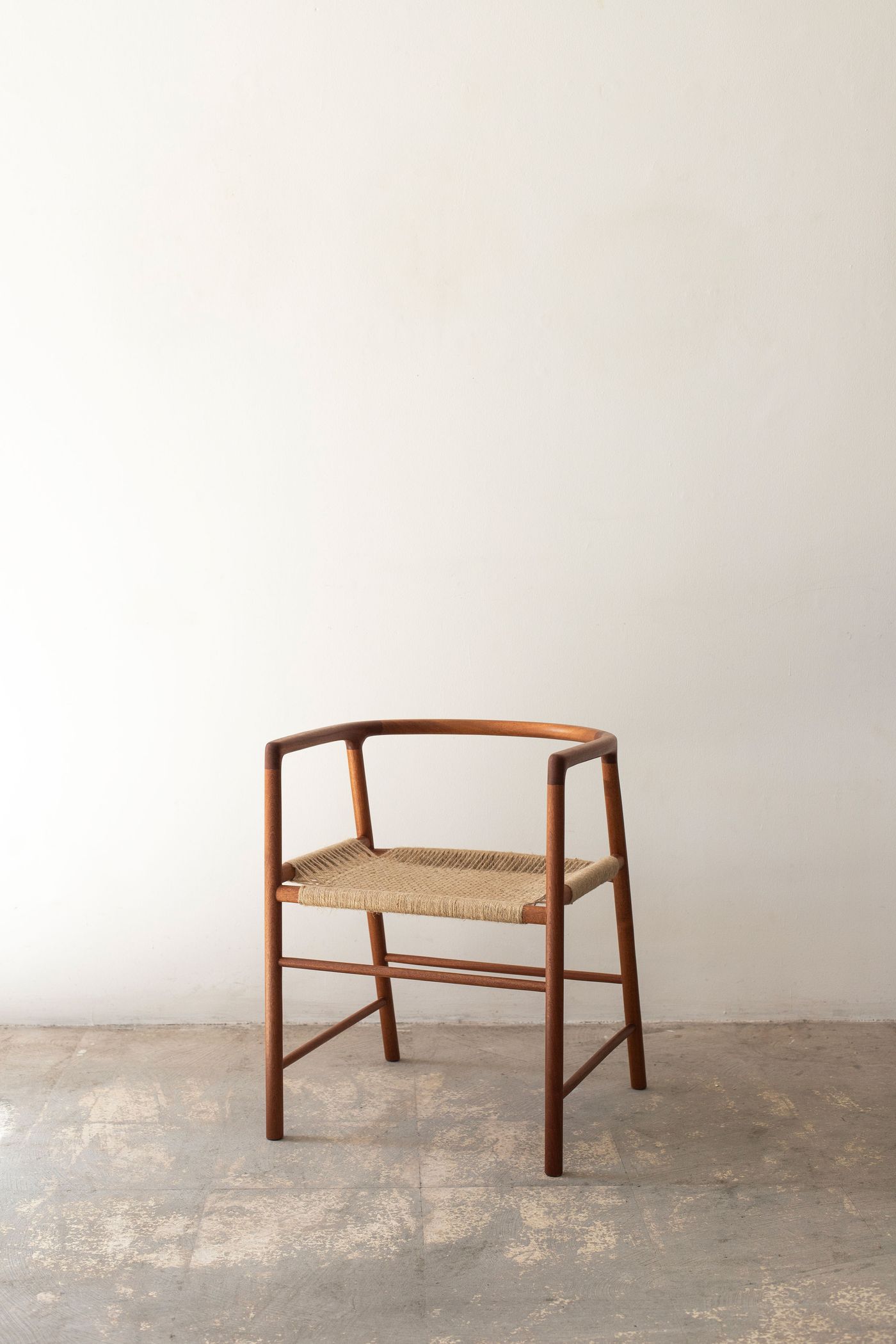 Midcentury-modern armchair exemplifies timeless Scandinavian design with its curved walnut frame and handwoven rush seat. The piece's graceful proportions and cross-braced structure demonstrate masterful woodworking, positioned against weathered concrete that emphasizes the chair's organic warmth and refined craftsmanship.