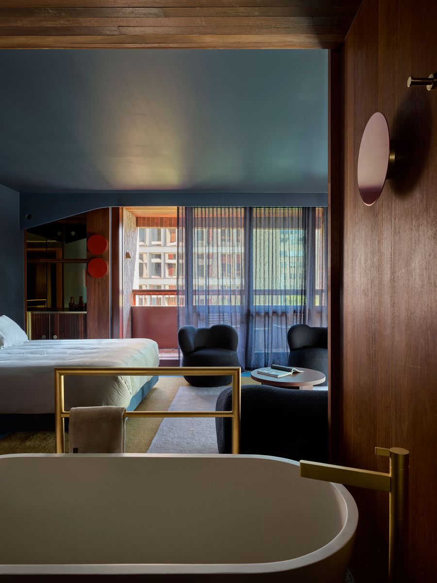 A stylish hotel room view from a wooden door, featuring a tub, bed, two black chairs, and large windows with sheer curtains. A stylish hotel room view from a wooden door, featuring a tub, bed, two black chairs, and large windows with sheer curtains.