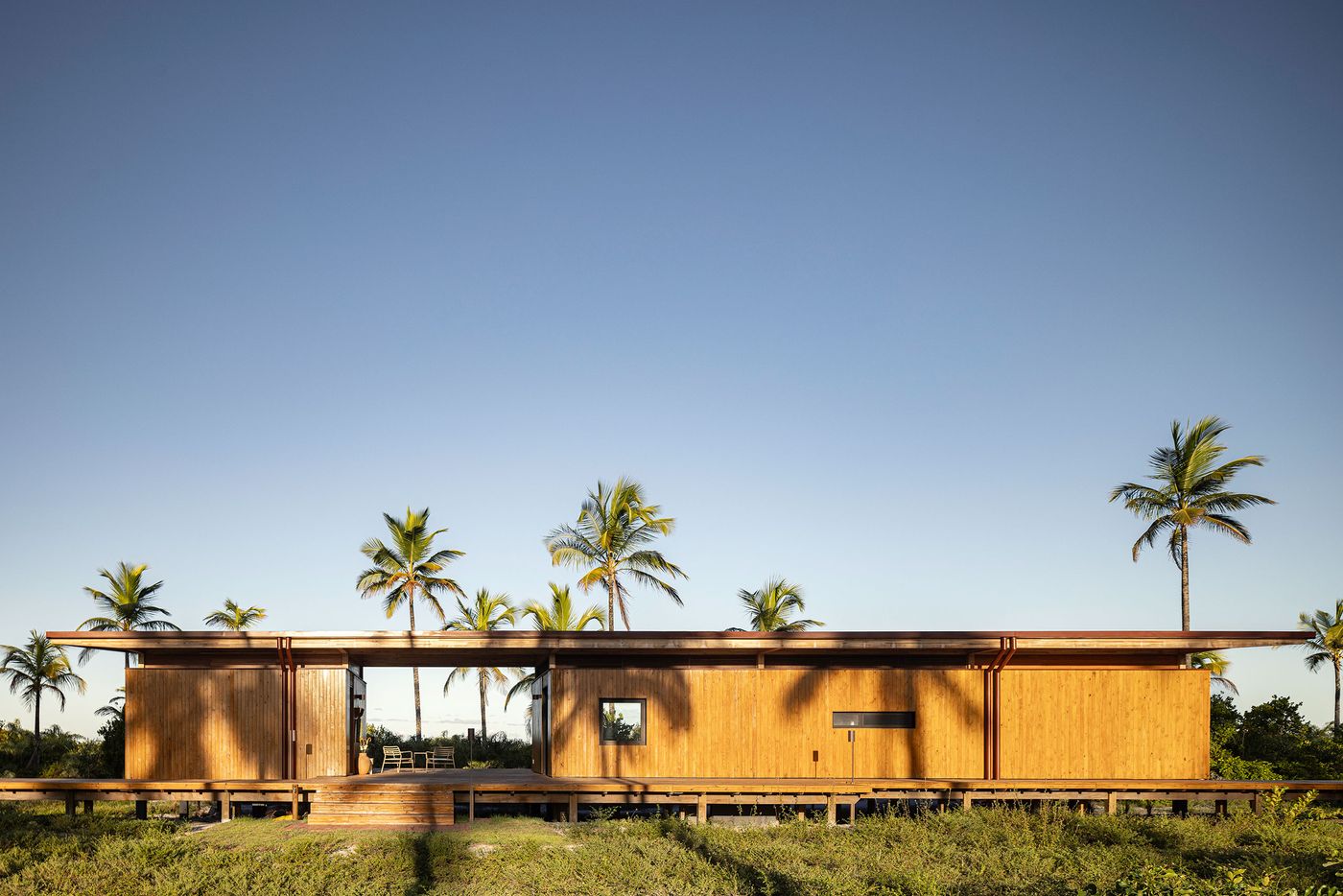 A modernist, low-slung structure with warm wooden siding sits elevated on a deck amidst a tropical landscape. Its clean horizontal lines and expansive openings invite the surrounding palm trees and clear blue sky indoors, embodying a seamless integration with nature and a relaxed, open design. A modernist, low-slung structure with warm wooden siding sits elevated on a deck amidst a tropical landscape. Its clean horizontal lines and expansive openings invite the surrounding palm trees and clear blue sky indoors, embodying a seamless integration with nature and a relaxed, open design.