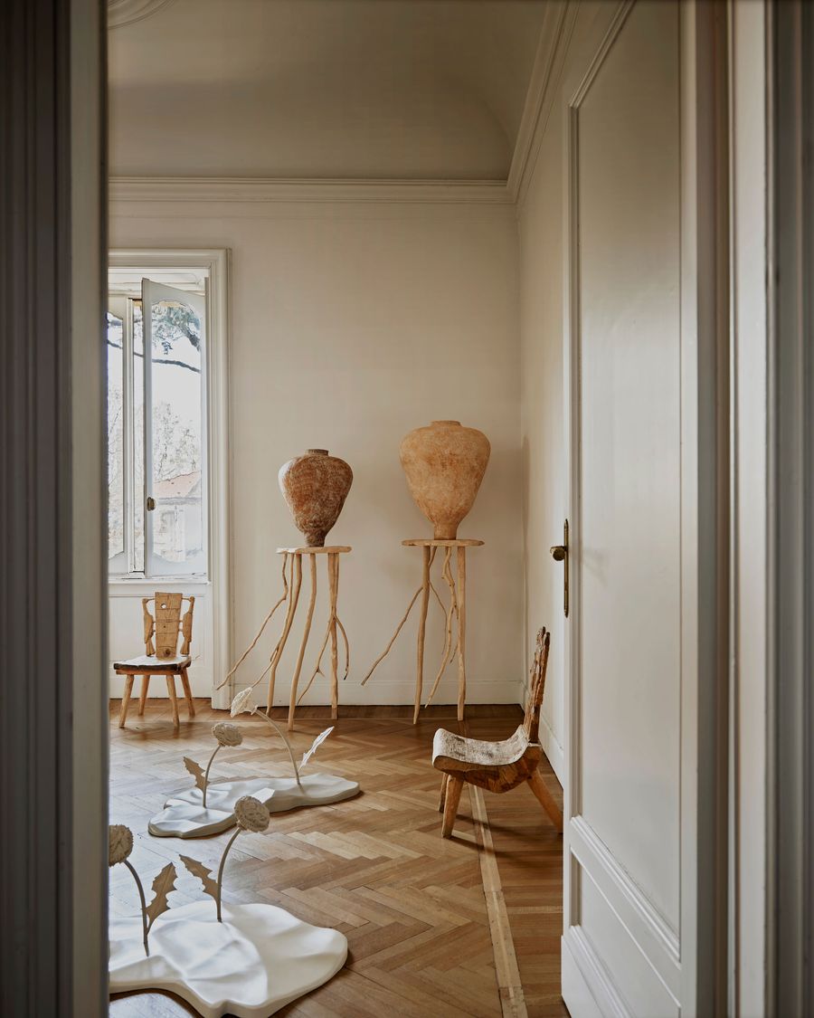 A minimalistic room featuring two tall clay vases, unique furniture, and artistic sculptures on a polished wood floor. Natural light streams in.