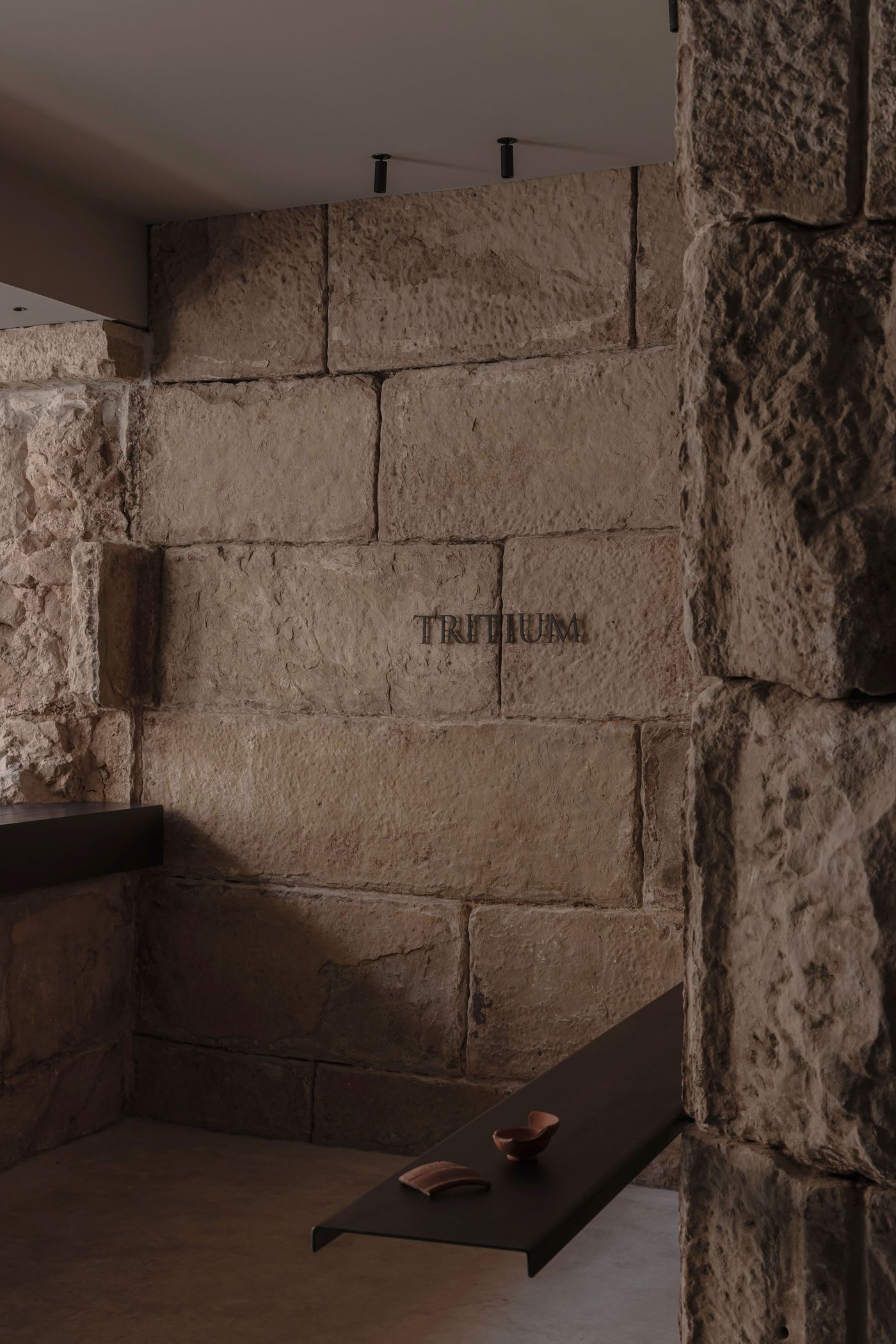 Interior view featuring rustic stone walls, a dark shelf with clay objects, and the word "Tritium" elegantly engraved on the wall.