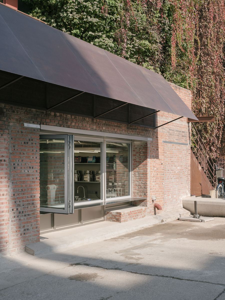 A sturdy brick building, part of the complex, is partially enveloped by vibrant green and reddish-brown foliage, blending industrial heritage with biophilic elements. A broad, dark awning extends over a large window, which opens outward, revealing a functional interior space with shelves and a sink, suggesting a potential cafe or workshop area. A sturdy brick building, part of the complex, is partially enveloped by vibrant green and reddish-brown foliage, blending industrial heritage with biophilic elements. A broad, dark awning extends over a large window, which opens outward, revealing a functional interior space with shelves and a sink, suggesting a potential cafe or workshop area.