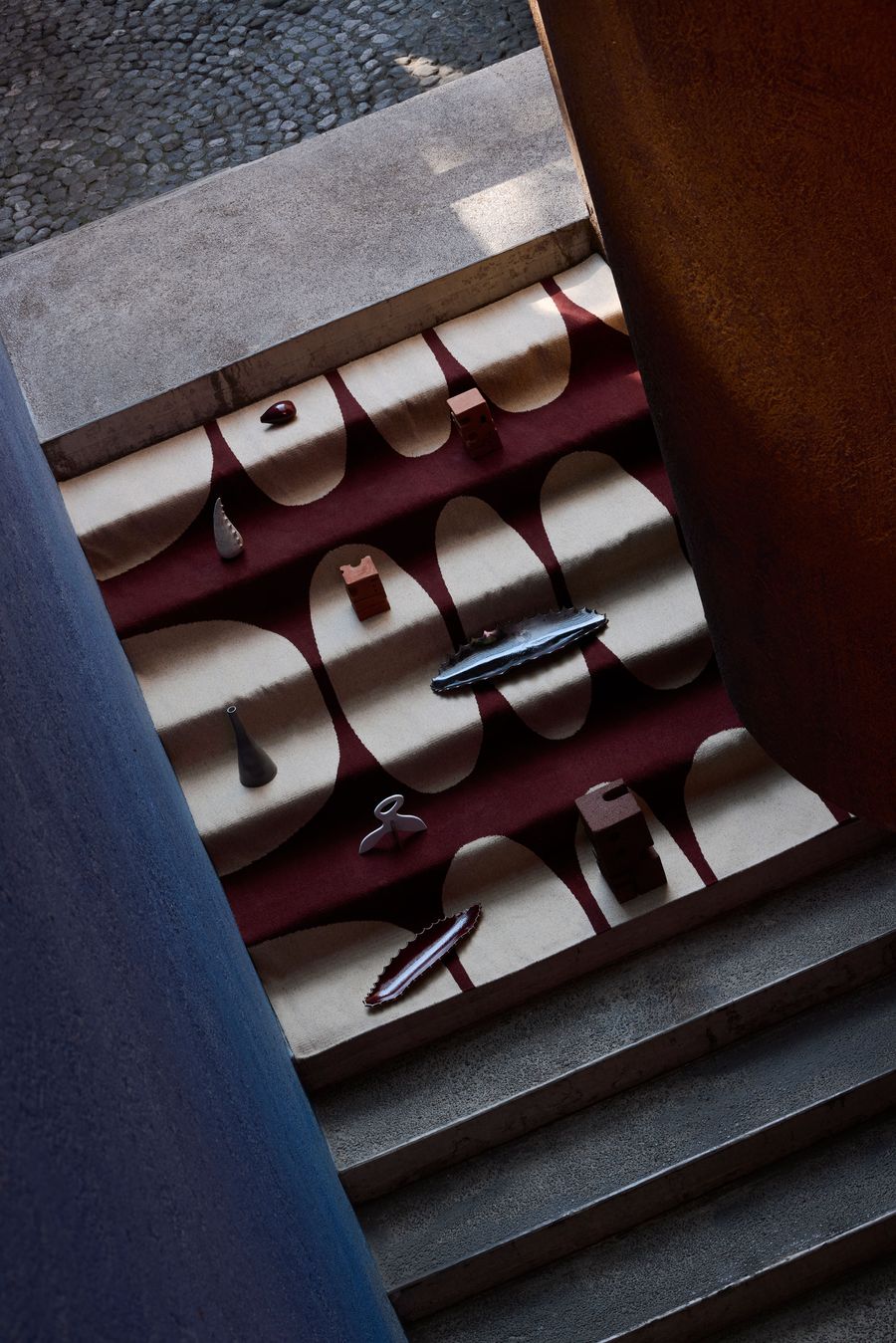 A top-down view of a patterned rug with various small sculptural objects arranged on it, set within a modern architectural space.