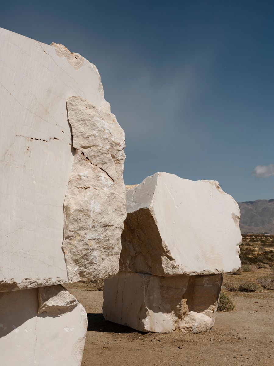 Two large, rough-hewn white marble blocks stand on a sandy landscape under a clear blue sky, with mountains in the distance. Two large, rough-hewn white marble blocks stand on a sandy landscape under a clear blue sky, with mountains in the distance.