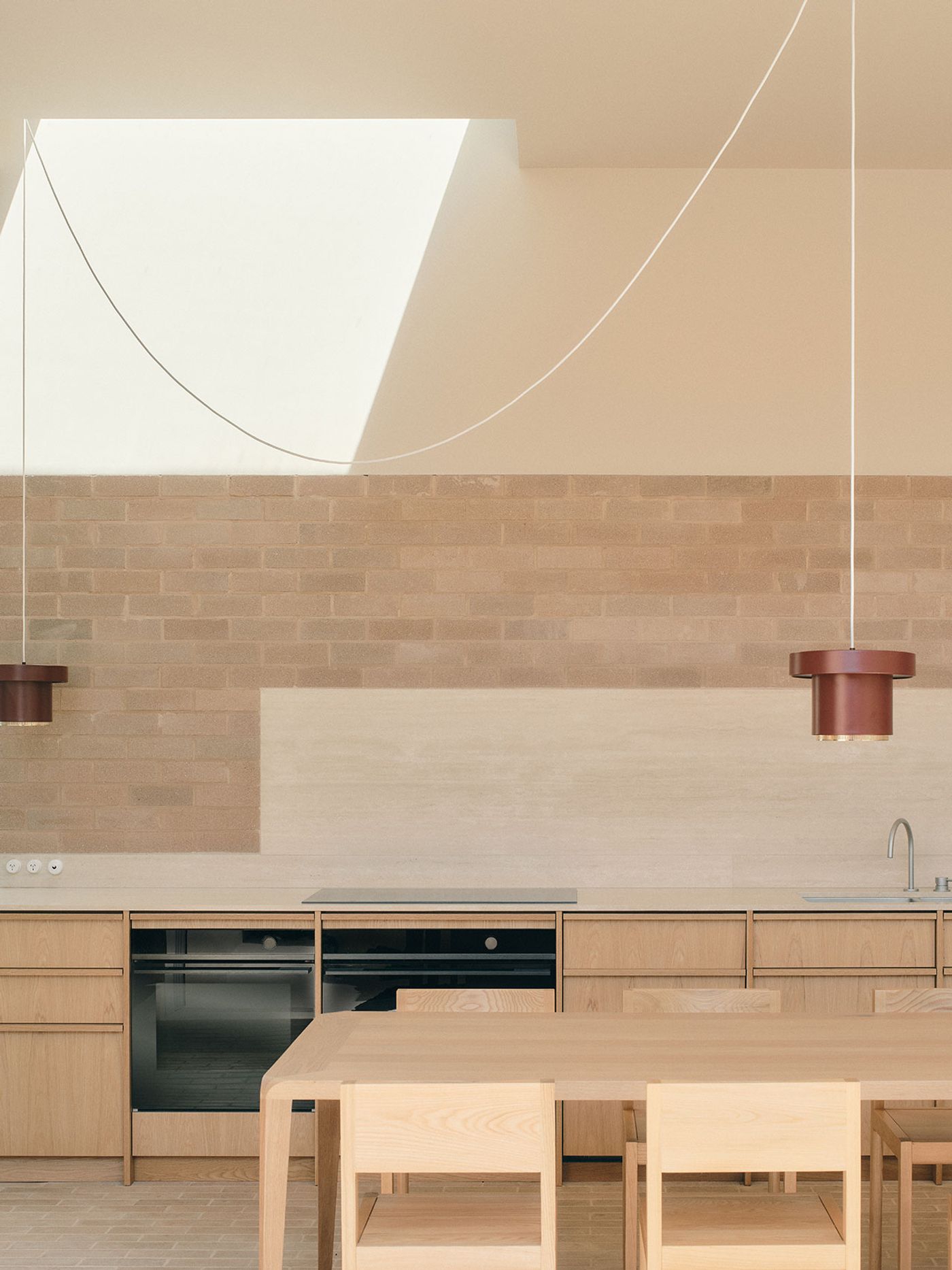 Modern kitchen featuring wooden cabinetry, minimalist design, a sleek countertop, and pendant lighting above a dining table. Natural light filters in. Modern kitchen featuring wooden cabinetry, minimalist design, a sleek countertop, and pendant lighting above a dining table. Natural light filters in.