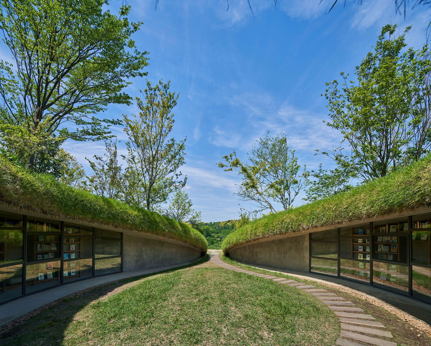 A green-roofed building with large glass windows sits in a grassy landscape under a clear blue sky, surrounded by trees. A green-roofed building with large glass windows sits in a grassy landscape under a clear blue sky, surrounded by trees.