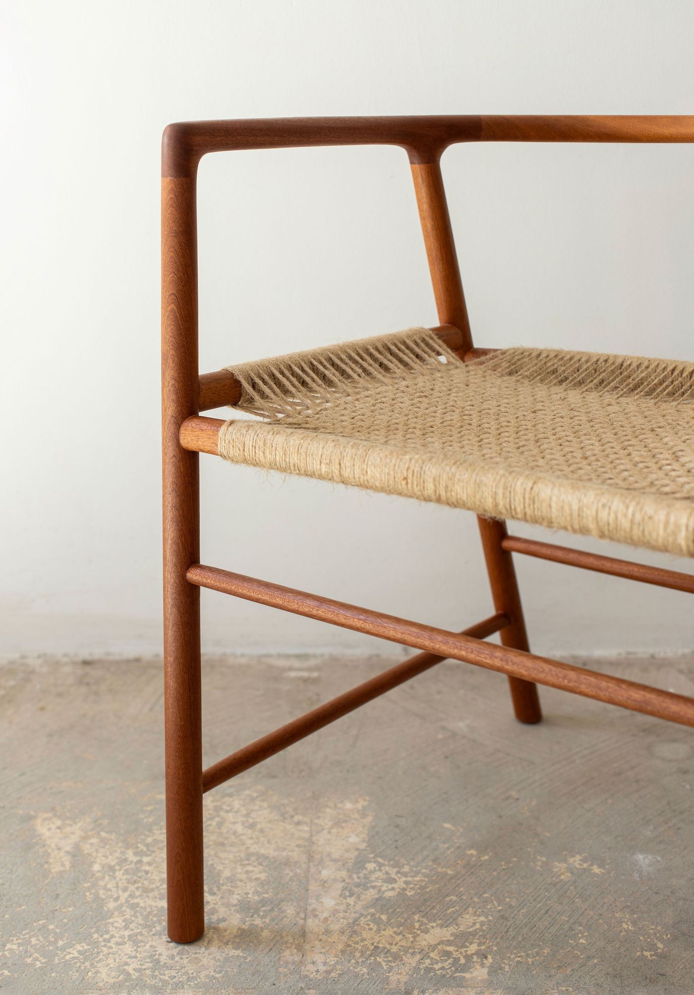 A close up of a chair reveals the intersection of wood and weave, where smooth walnut joinery meets intricate rush cordage. The chair's sculptural frame creates geometric shadows while the natural fiber seat introduces organic texture, showcasing the dialogue between structured design and handcrafted elements.